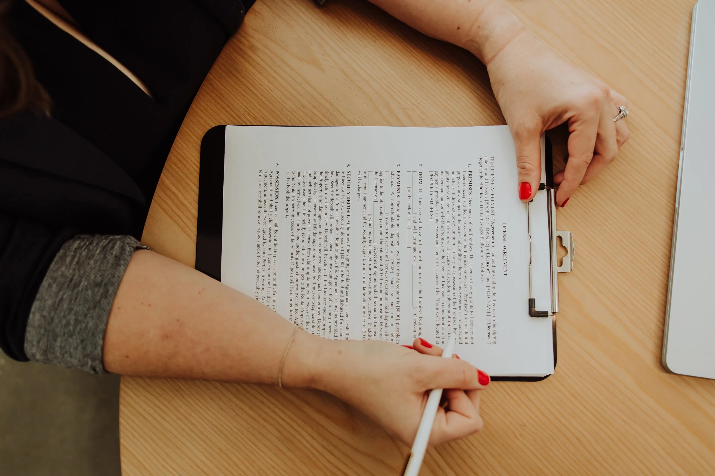 A person with red nail polish and a ring is holding a pen and reviewing a document on a clipboard at a wooden desk. The person is wearing a black jacket and has a gray sleeve rolled up.