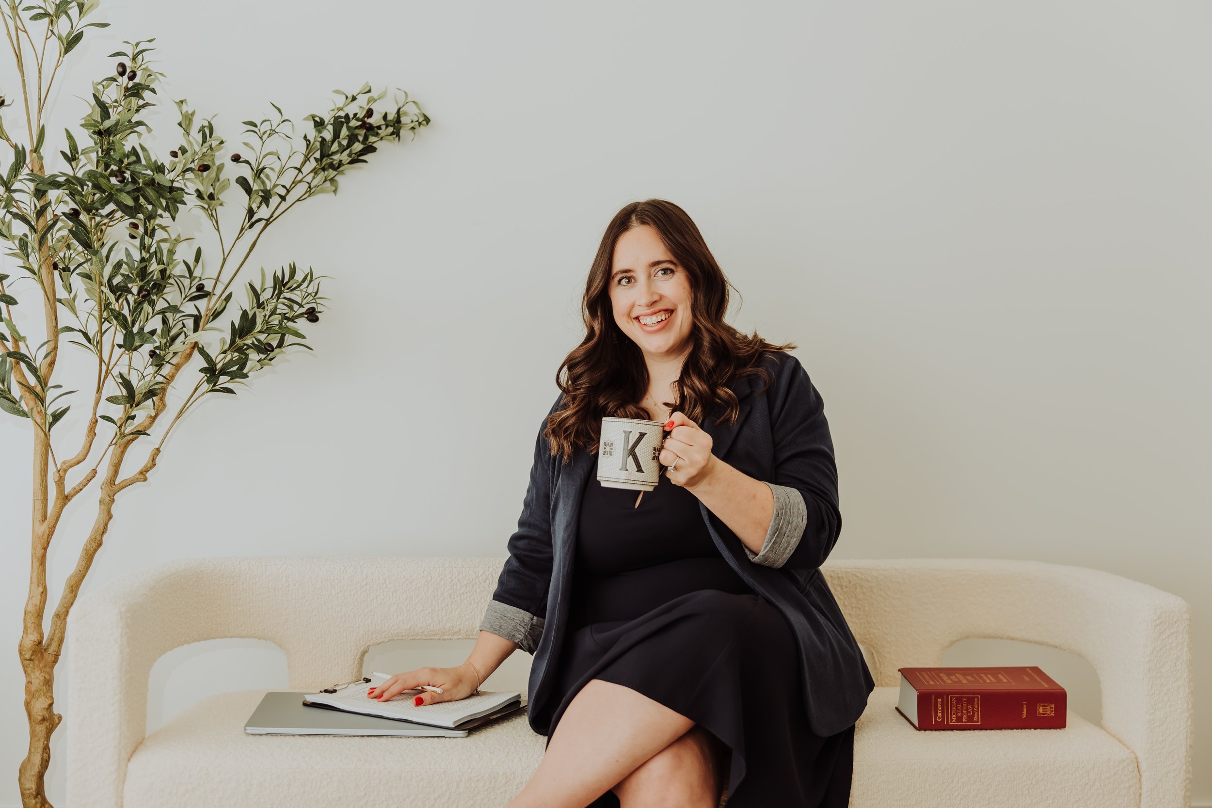 A woman with long brown hair, wearing a black dress and black blazer, sitting on a cream-colored couch, holding a mug with the letter 'K' on it, smiling at the camera. There is a computer and a red book on the couch beside her, and a green houseplant to her left.