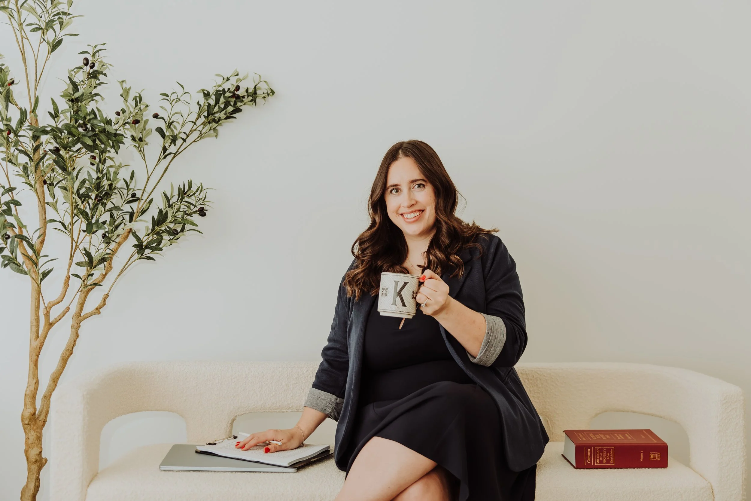 Woman sitting on a beige couch, holding a mug with the letter 'K', smiling, with a large book and a notebook nearby, and a tall green plant beside her against a plain light-colored wall.