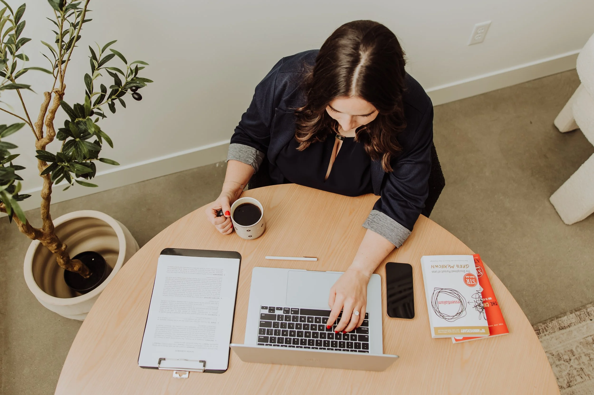 A woman sitting at a round table with a laptop, a phone, a clipboard with papers, a pen, a book, and a cup of coffee.
