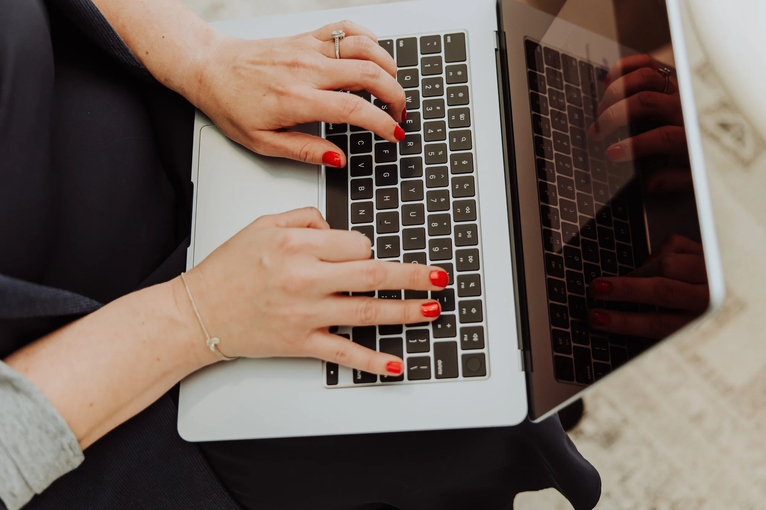 A person with red nail polish, wearing a ring and a bracelet, using both hands to type on a laptop keyboard.