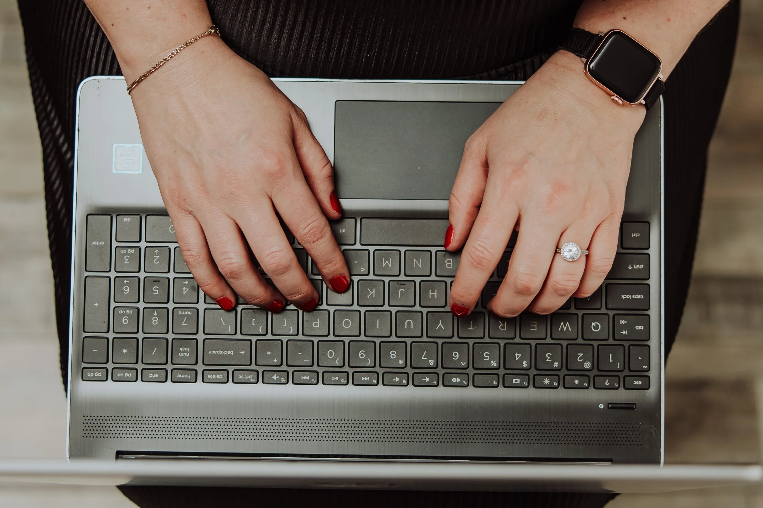 A person with painted red nails and a diamond ring on their left hand typing on a laptop keyboard.