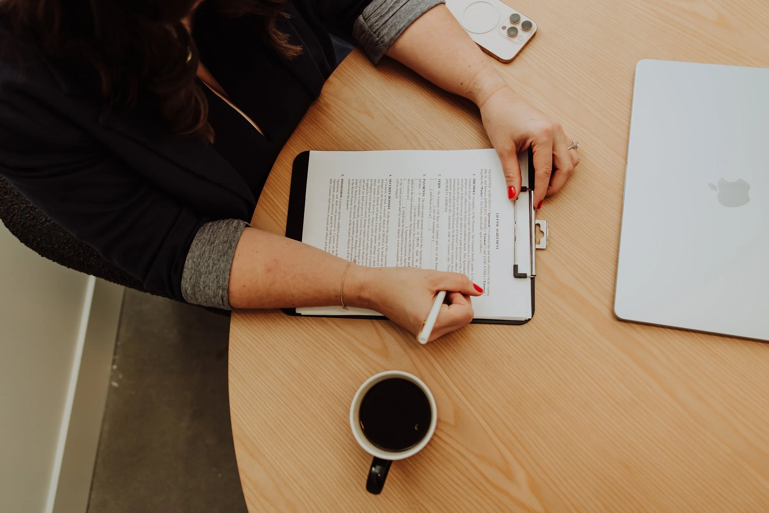 Person writing on a clipboard with a document, with a cup of coffee, a laptop, and a smartphone on a wooden desk.