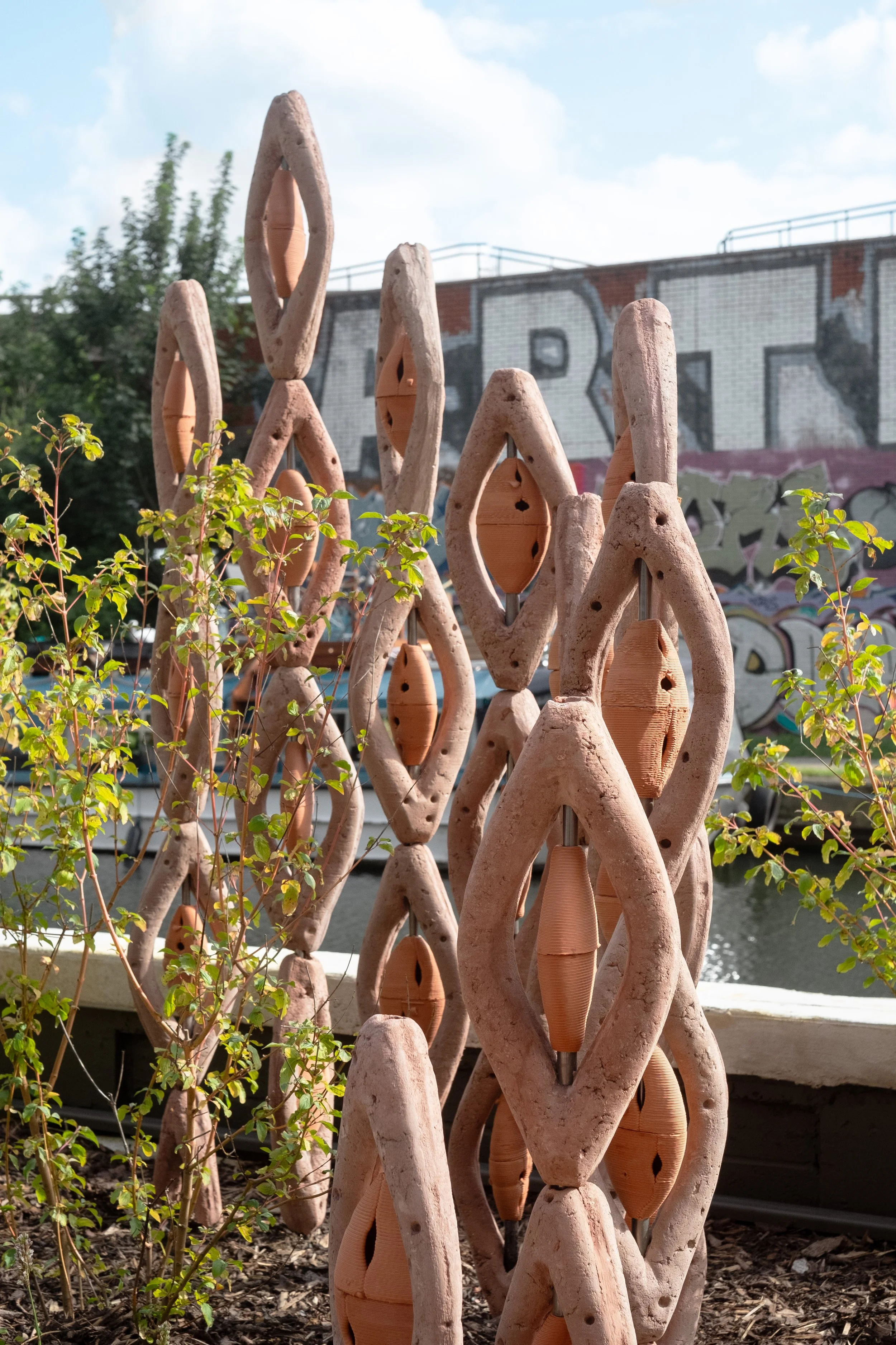 Outdoor sculpture made of interconnected, textured, clay-colored shapes with wooden and metal components, near a small shrub, with graffiti wall and cityscape in the background.