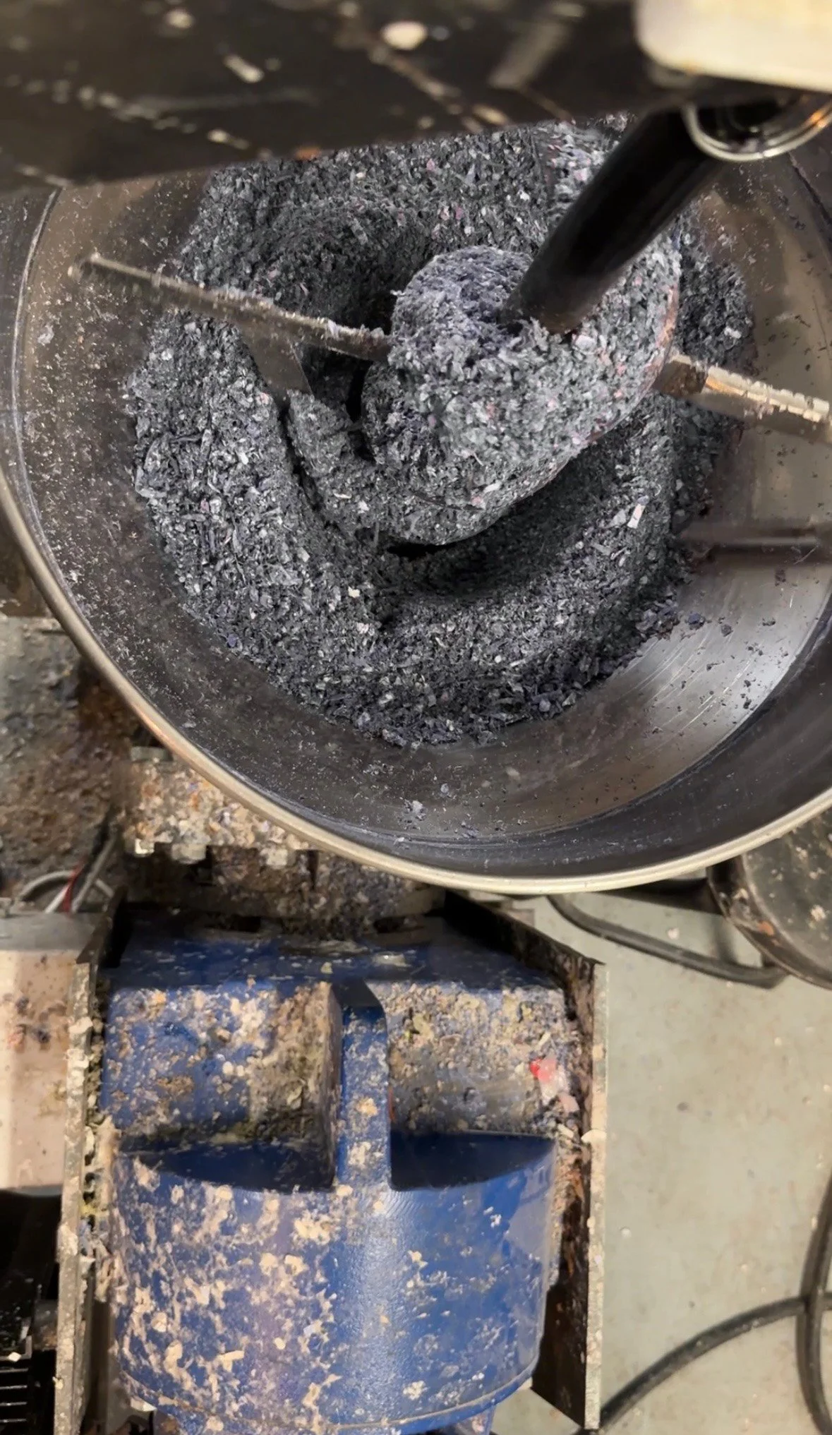 Close-up of an industrial machine blending black ashes or charcoal in a metal barrel, with a blue container and dust on the surrounding area.