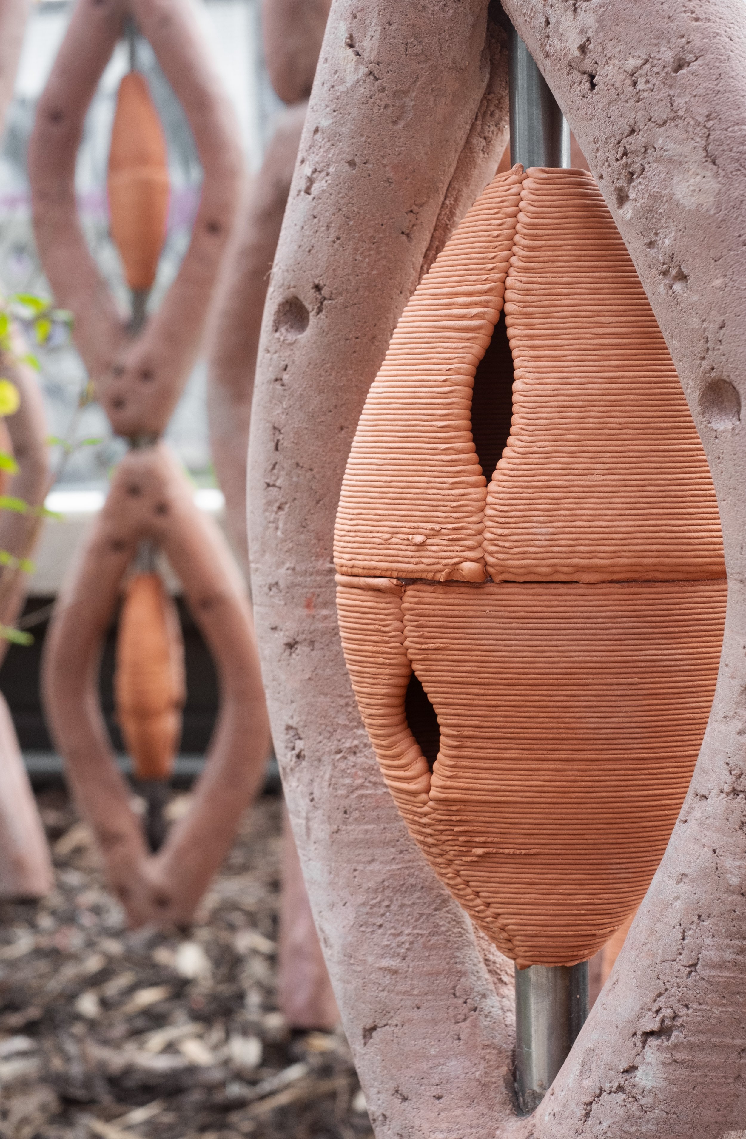 Close-up of a terracotta sculpture with ribbed texture, mounted on a metal rod, with similar sculptures blurred in the background.