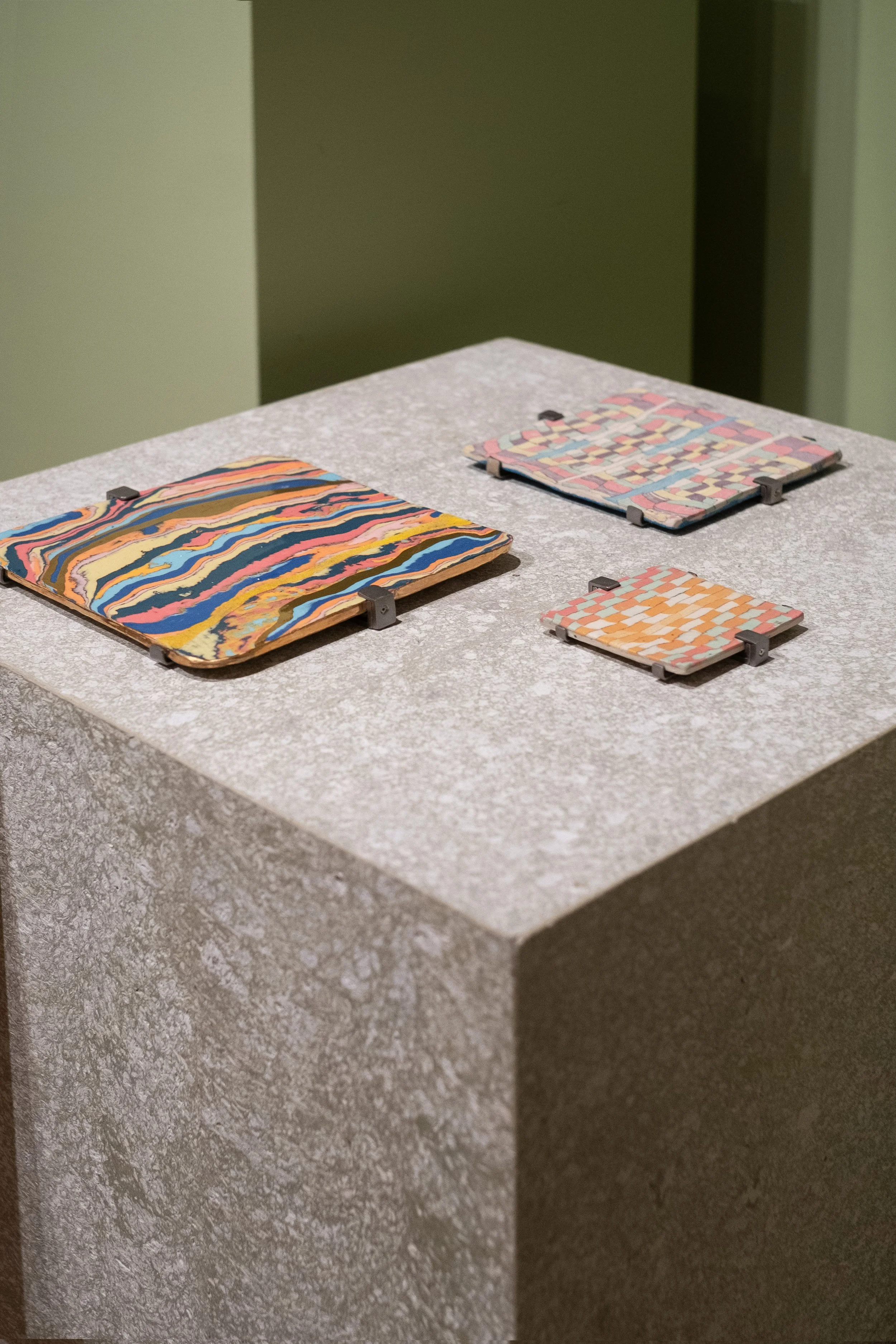 Three colorful patterned ceramic tiles displayed on a gray pedestal in a gallery setting.