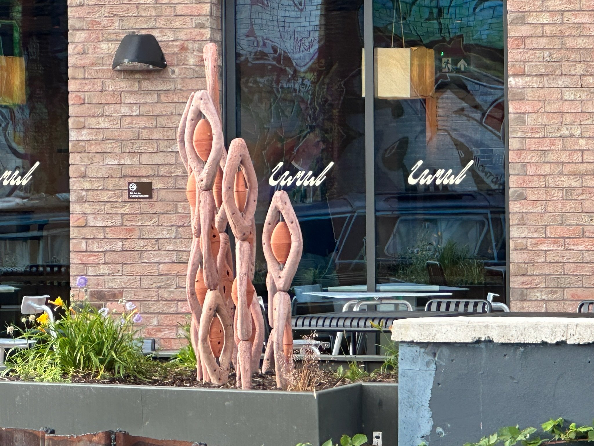 Decorative sculpture made of intertwined wooden and orange materials outside a building with brick walls and large glass windows.