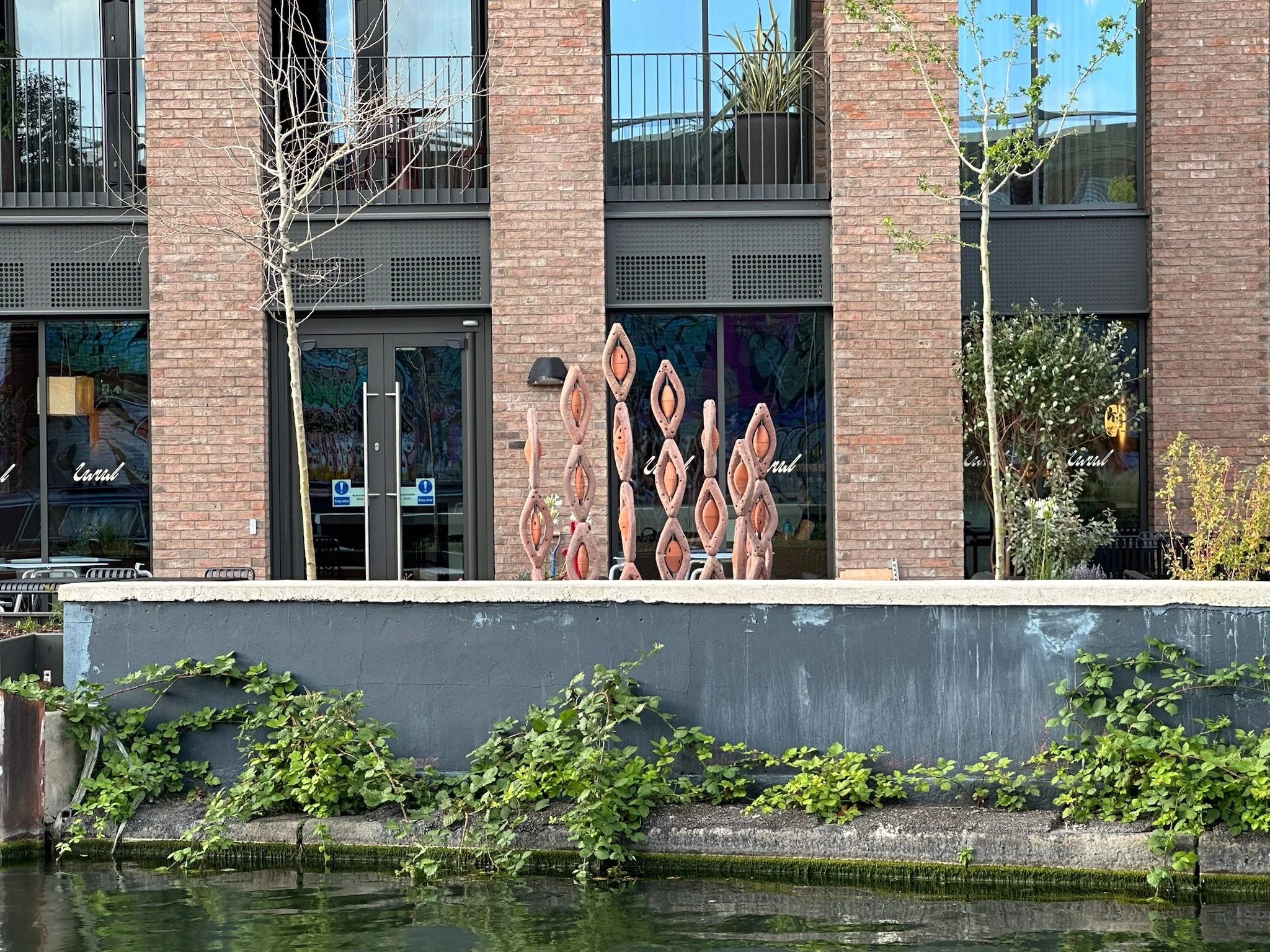 Modern brick building with large glass windows and plants on balconies, graffiti art on interior wall visible through glass, and outdoor sculptures resembling elongated, eye-shaped forms placed in front of large glass doors, surrounded by greenery an