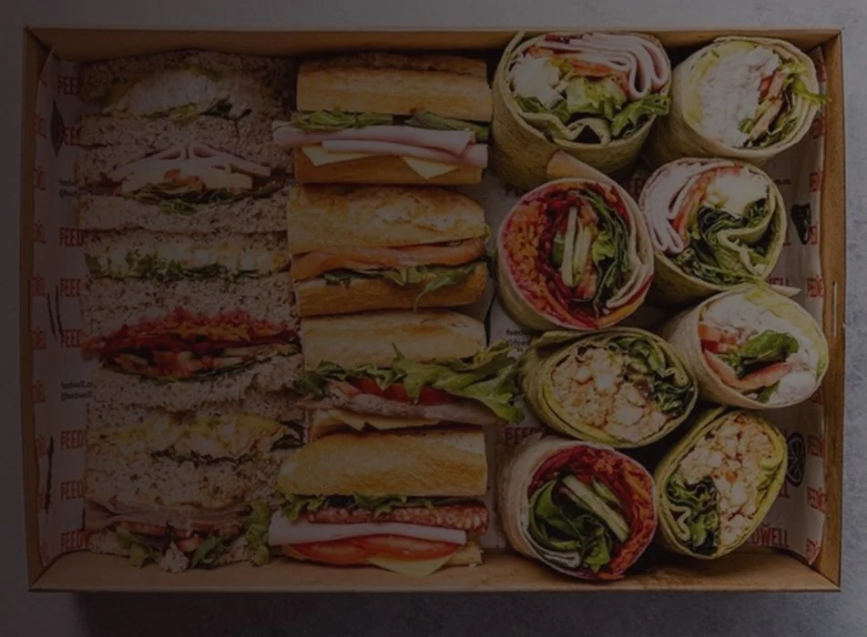 Assorted sandwiches and wraps in a box, including club sandwiches and veggie wraps.