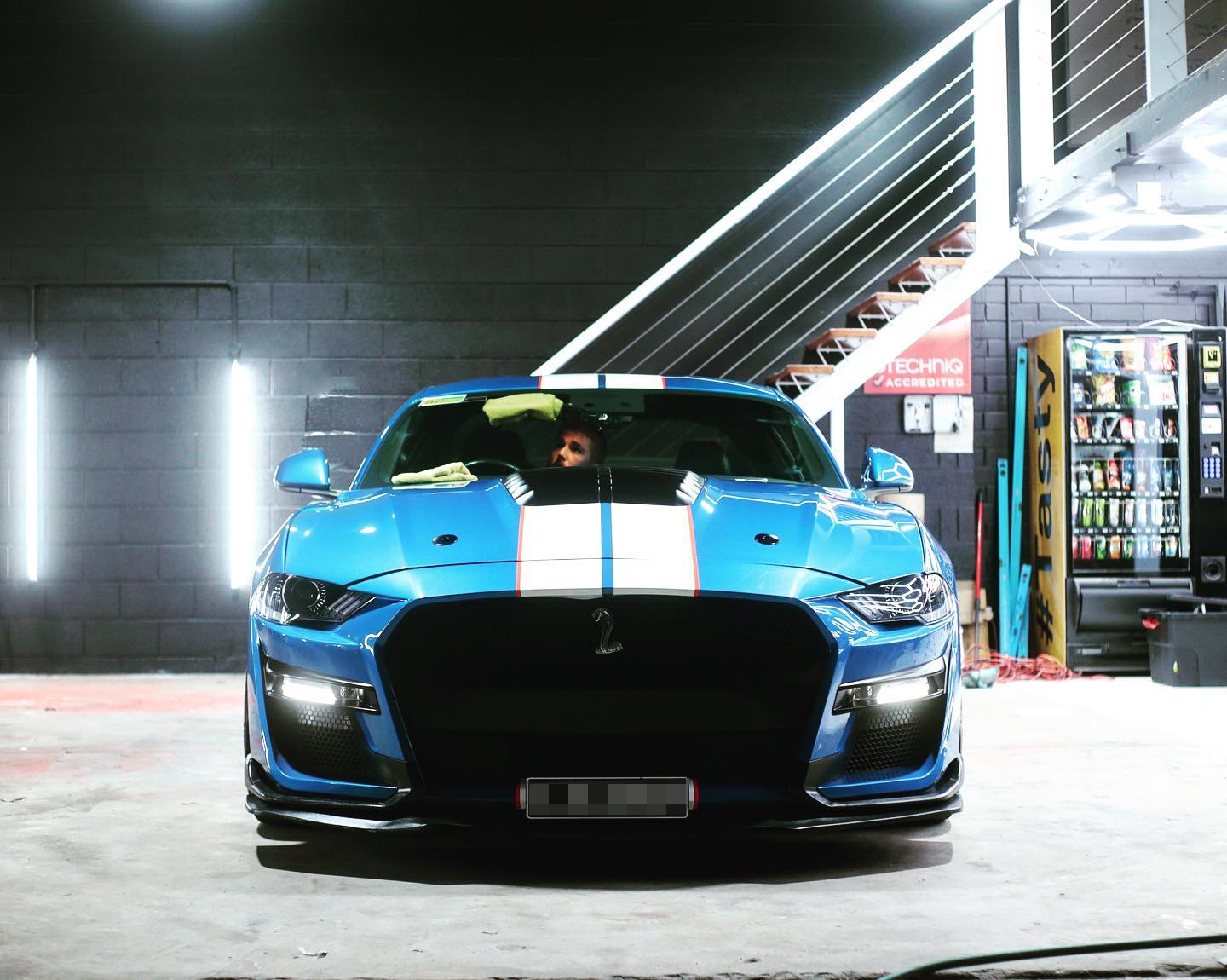 A blue sports car parked inside a garage with cleaning cloths on the windshield, vending machine to the right, and a staircase in the background.