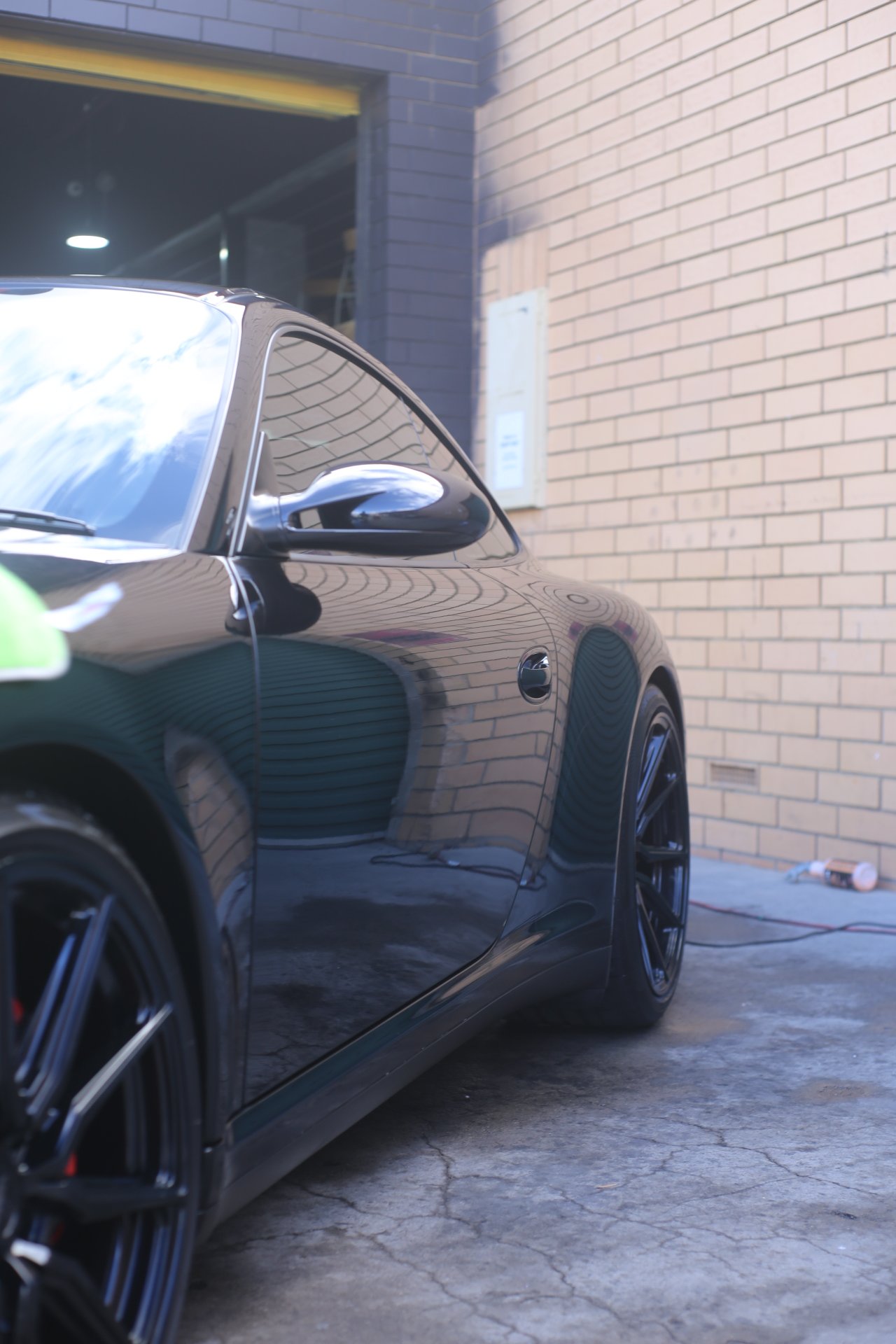 Black luxury sports car parked next to a brick wall, with a reflection of the sky and clouds on its shiny surface, and a power cord connected to it near the ground.