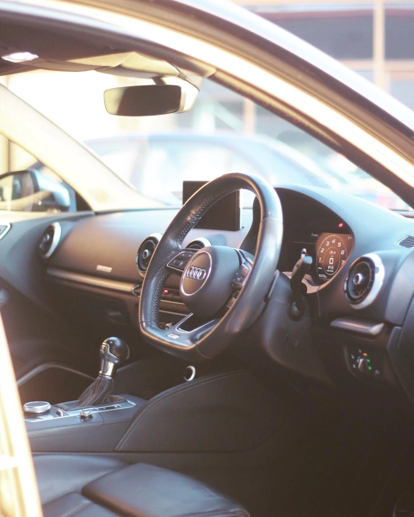 Interior of a car with a black steering wheel, dashboard, and gear shift lever. The dashboard displays a speedometer and other gauges.