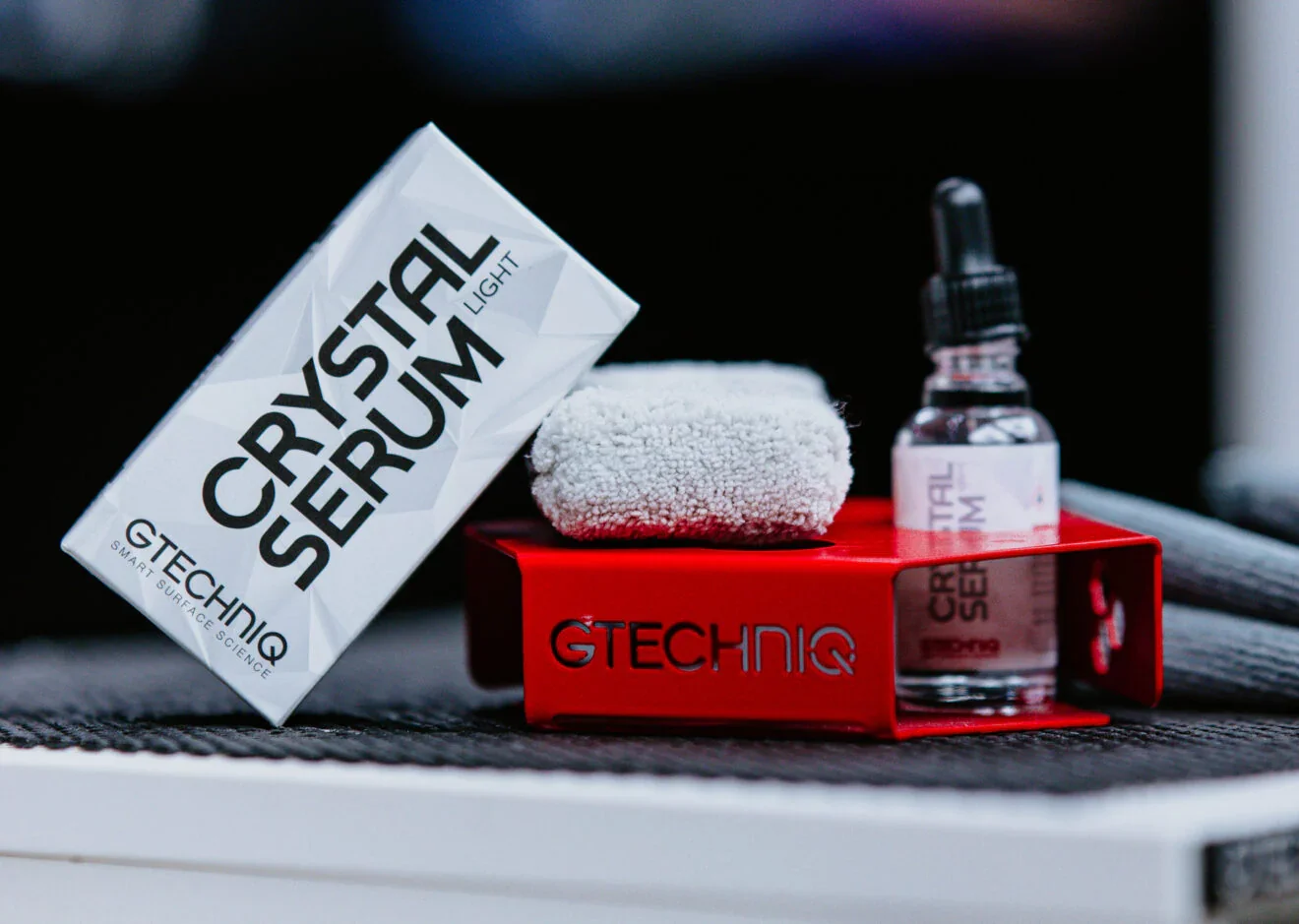 A cleaning kit with a bottle of ceramic sealer, a microfibre cloth, and a box labeled 'Ceramic Sealer Light' placed on a black surface with a red Gtechniq stand.