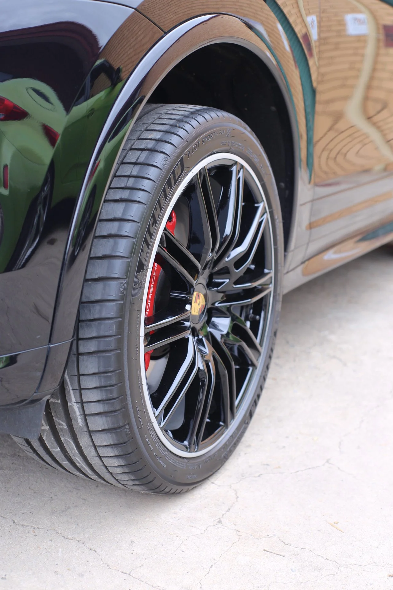 Close-up of a black luxury car's front tire and wheel, showing the Porsche emblem on the wheel hub and red brake calipers, parked on a concrete surface.