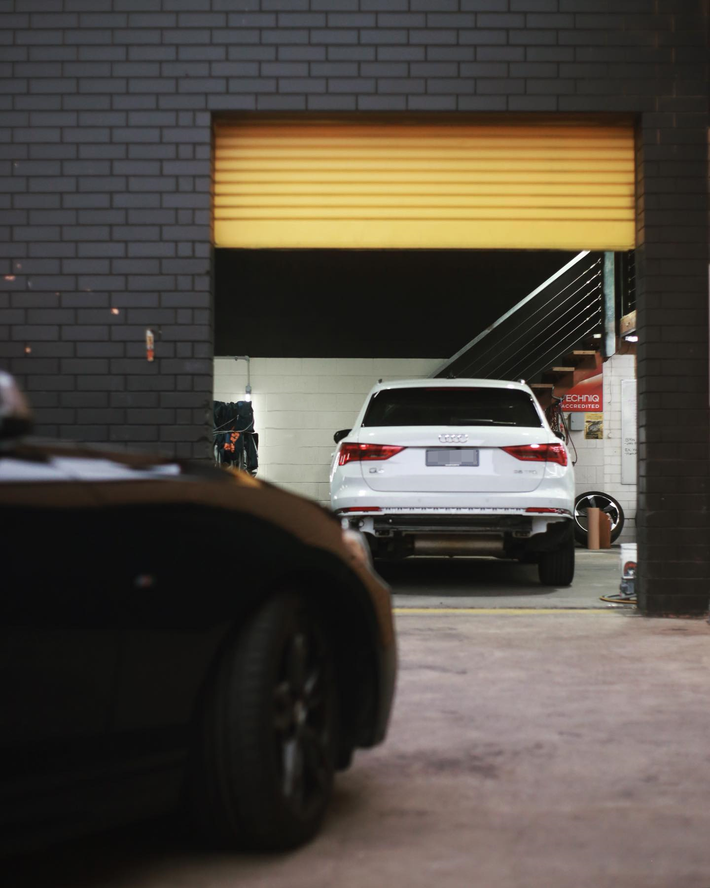 A white Audi SUV parked inside a garage with a partially open yellow roll-up door. The garage has black brick walls and visible stairs. The front of a black car is visible in the foreground.