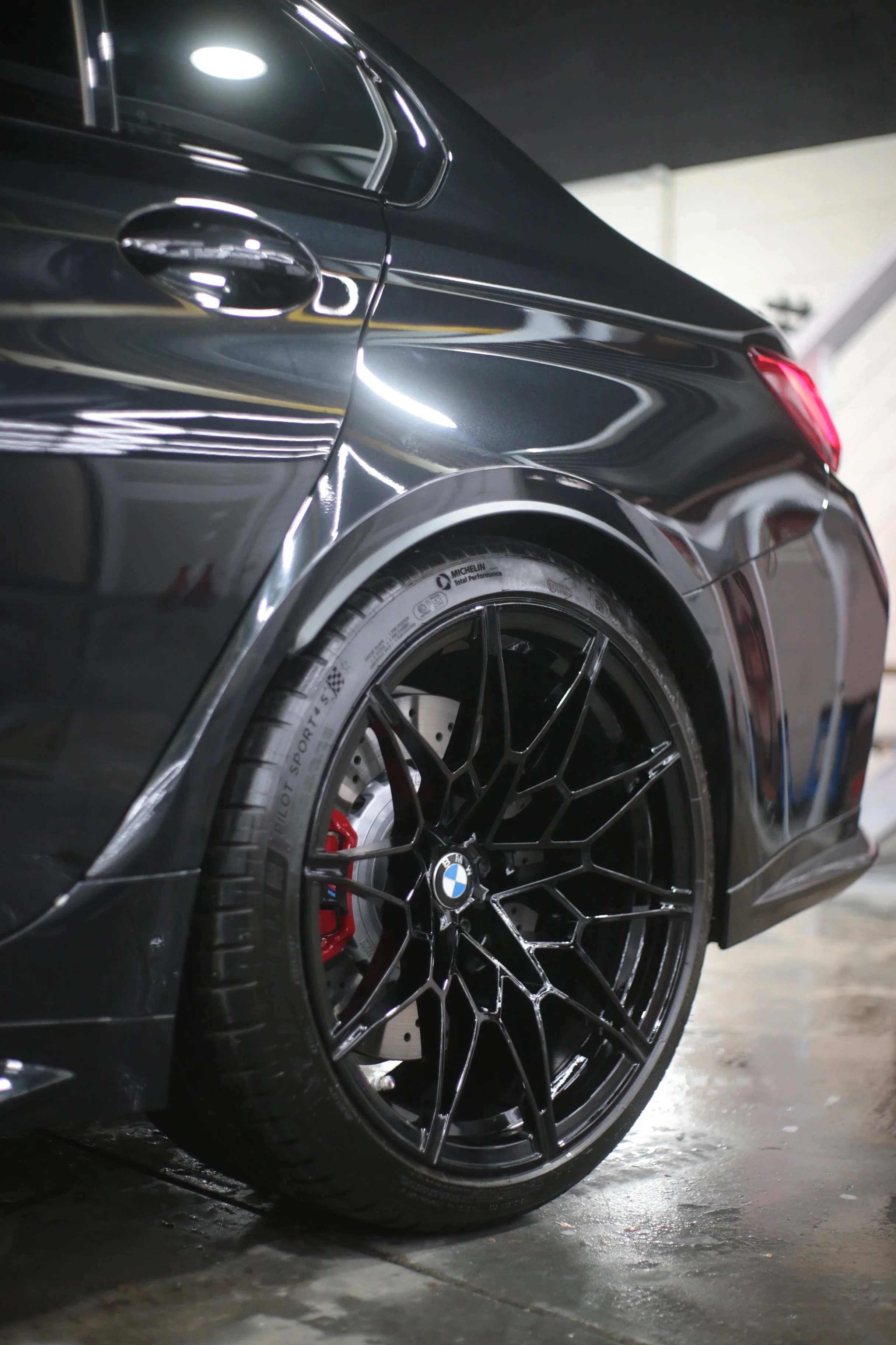 Close-up of the rear side of a black BMW sports car showing a detailed black alloy wheel, red brake caliper, tire, and part of the car's body with reflections, set in a garage with a concrete floor and overhead lighting.
