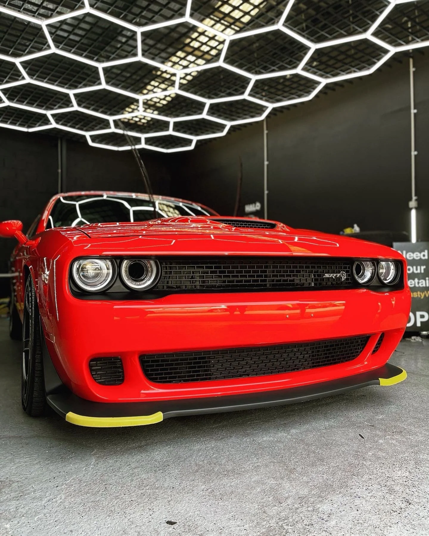 Red Dodge Challenger SRT sports car with black accents and yellow wheel protectors, parked indoors under a hexagonal light fixture.