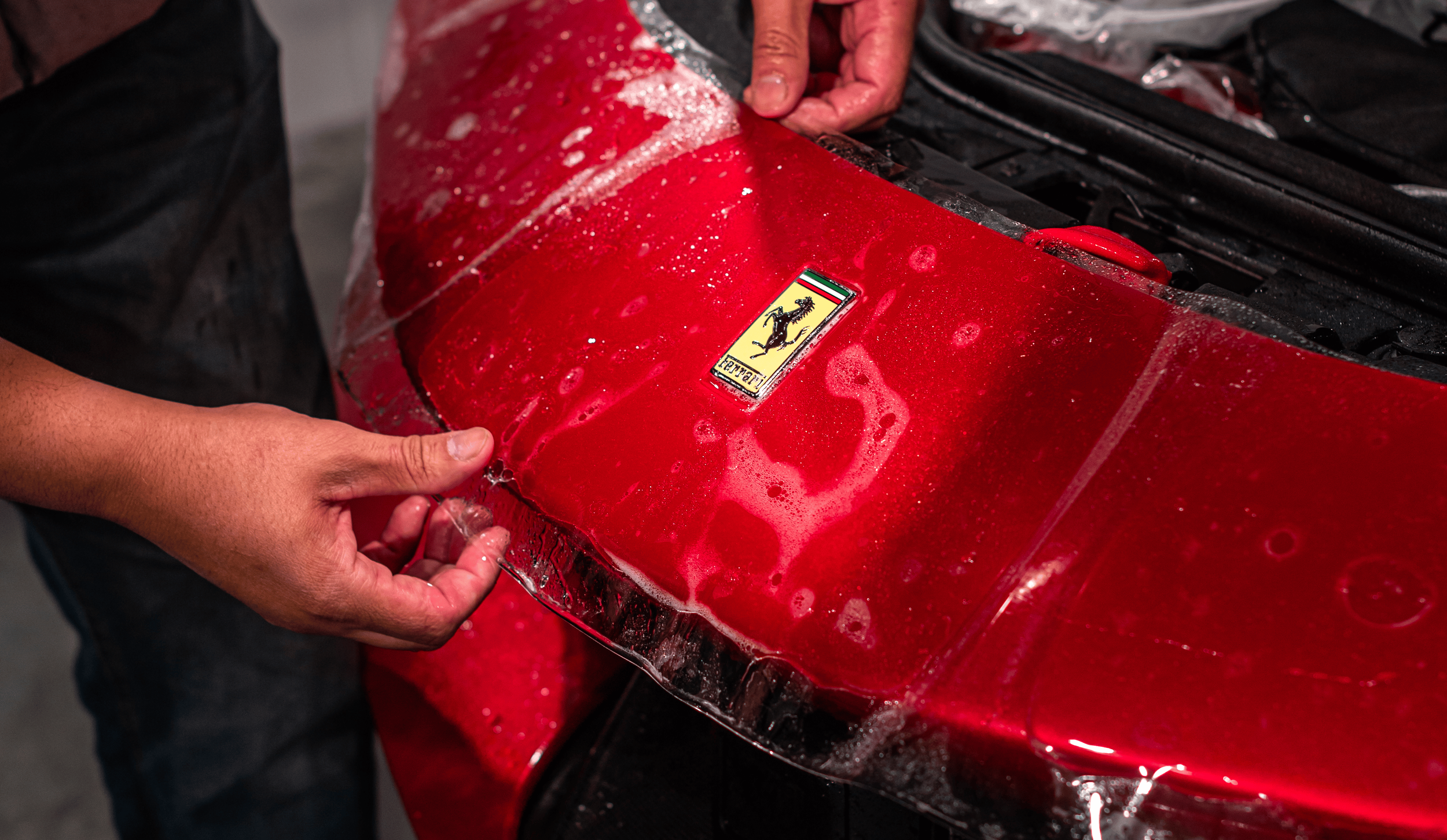 The image shows a person washing a red Ferrari car with soap and water, focusing on the car's front hood with the Ferrari logo.