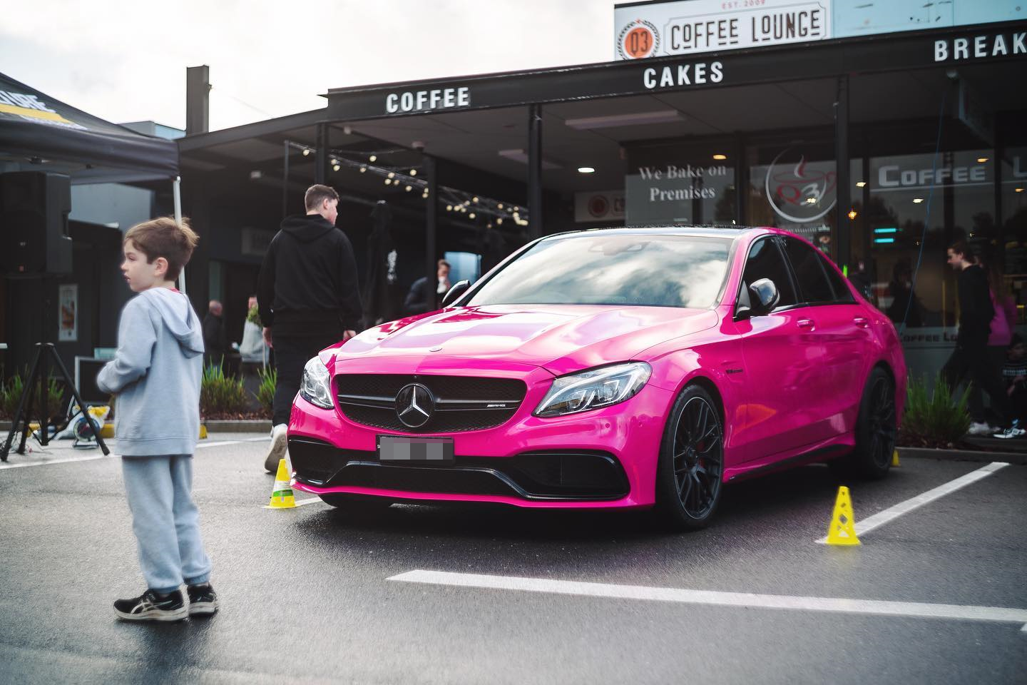 Pink Mercedes-Benz car parked outside a coffee shop with people around, including a boy in a gray hoodie in the foreground.