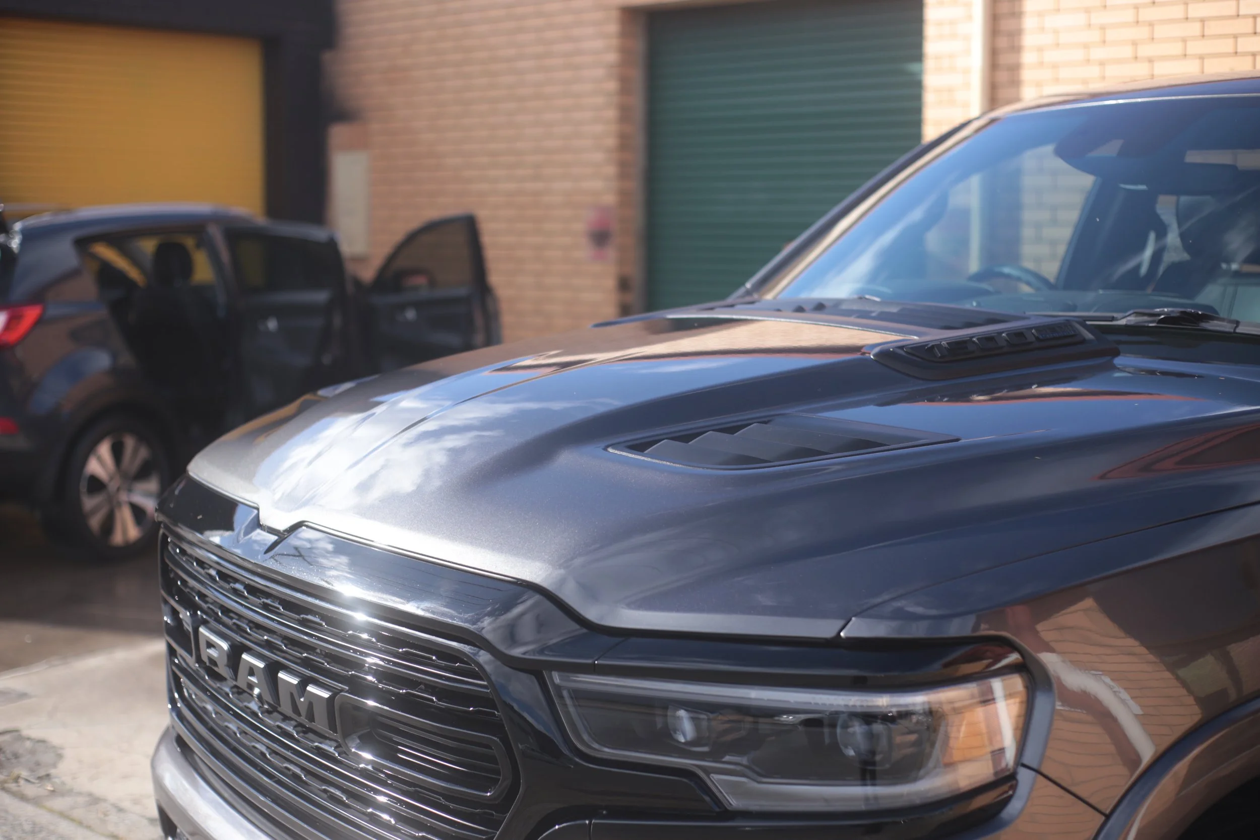 A silver RAM truck parked outdoors with another vehicle nearby and a brick building in the background.