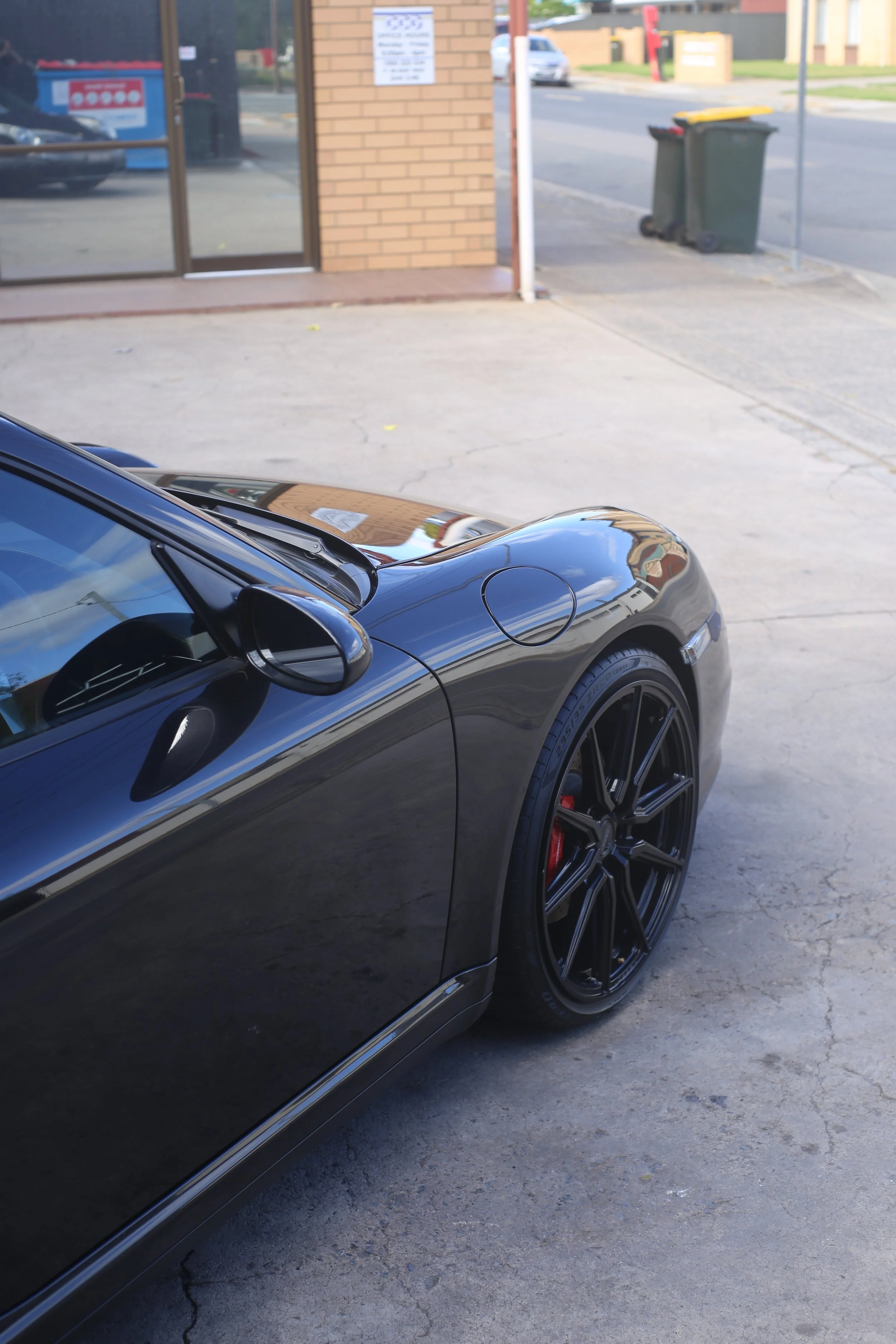 Close-up of the front part of a black sports car with custom black wheels parked outside a building, with a brick wall, a glass door, and trash bins in the background.