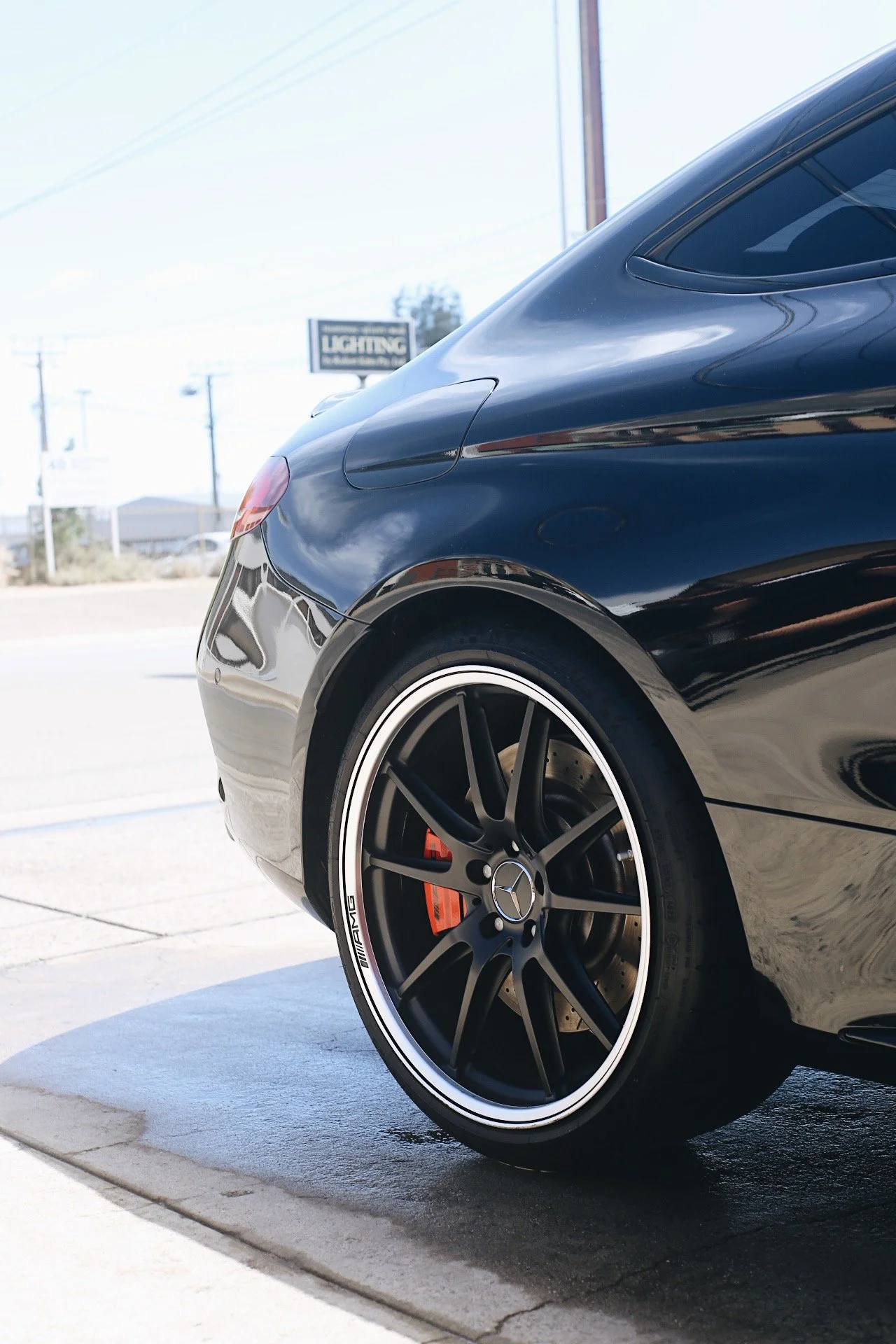 Close-up of the rear section of a black luxury car with alloy wheels and tinted windows, parked on a concrete surface with a background of power lines and buildings.