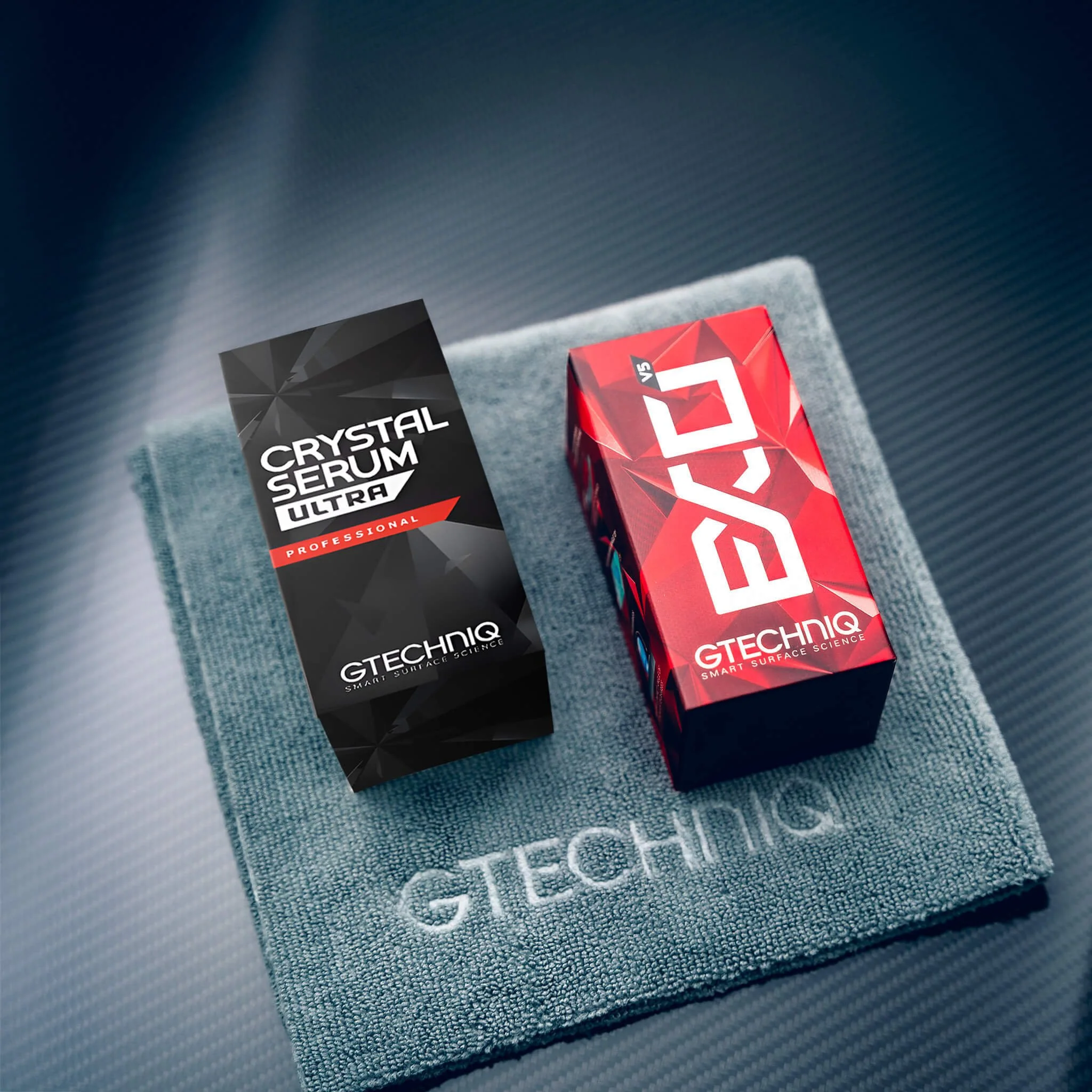 Close-up of a gray textured towel with the word 'TECHIQ' embroidered on it, and two boxes of surface science products, one labeled 'CRYSTAL SERUM ULTRA' and the other labeled 'GTECHIQ' with red and black geometric designs.