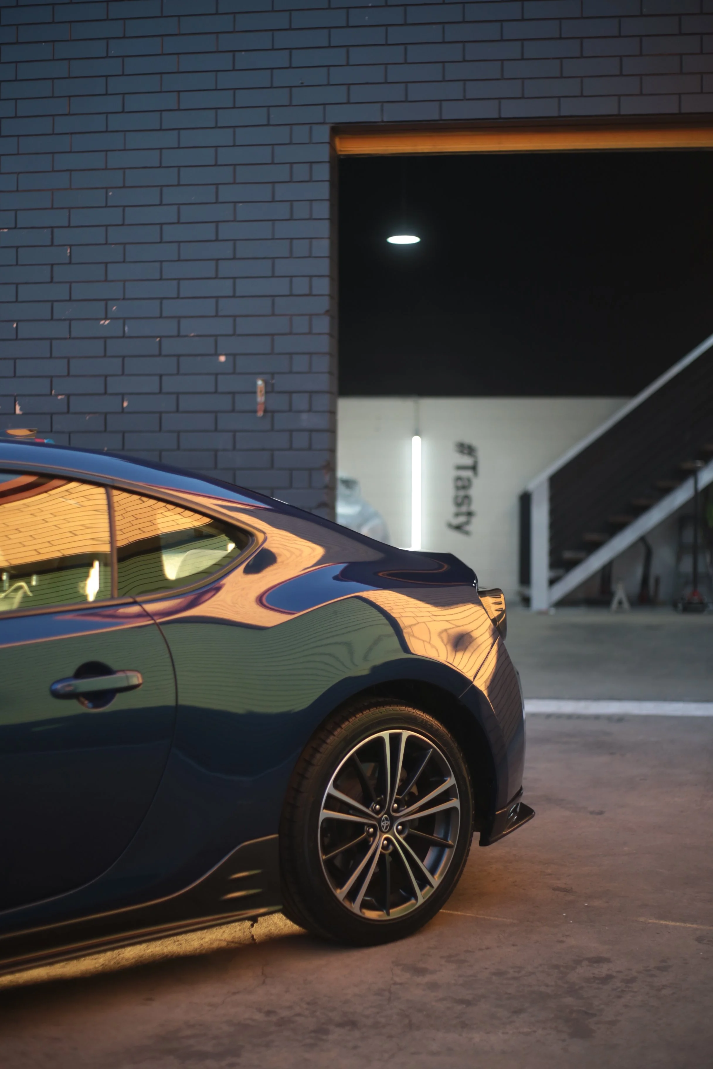 Close-up of a black modern car parked in front of a building, with a brick wall and staircase in the background, illuminated by ambient lighting.