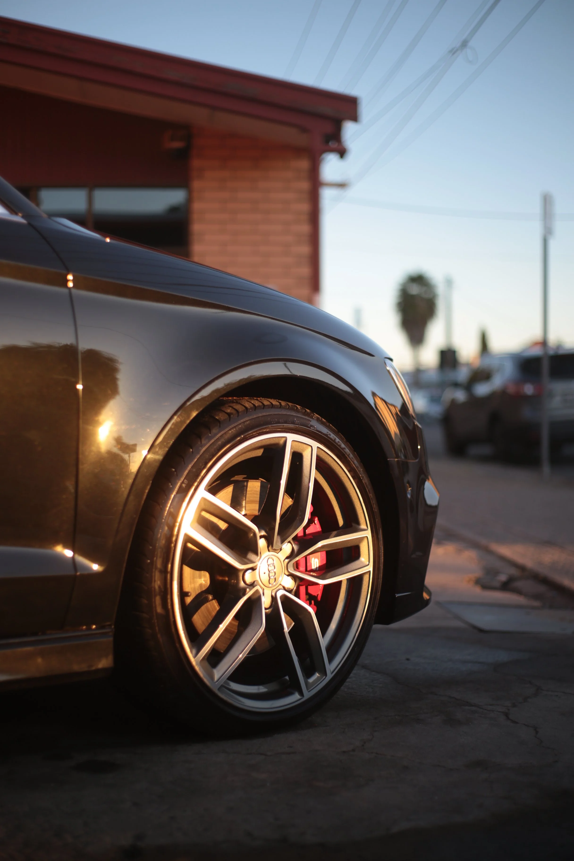 Close-up of a black luxury car's front wheel with red brake caliper, parked on a city street during sunset, with a brick building, power lines, and a palm tree in the background.