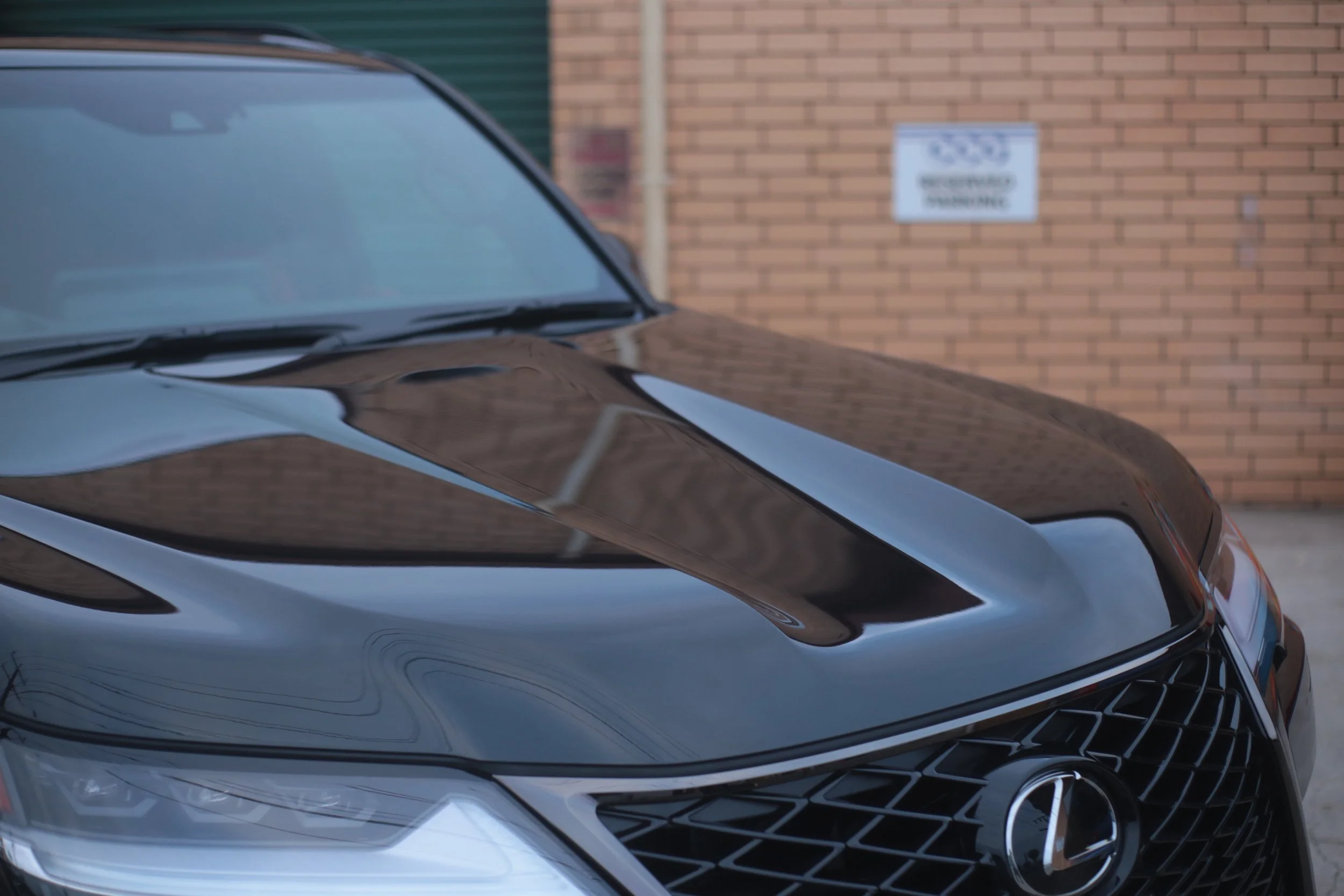 Close-up of the front end of a shiny black luxury car, with a brick wall and a sign in the background.