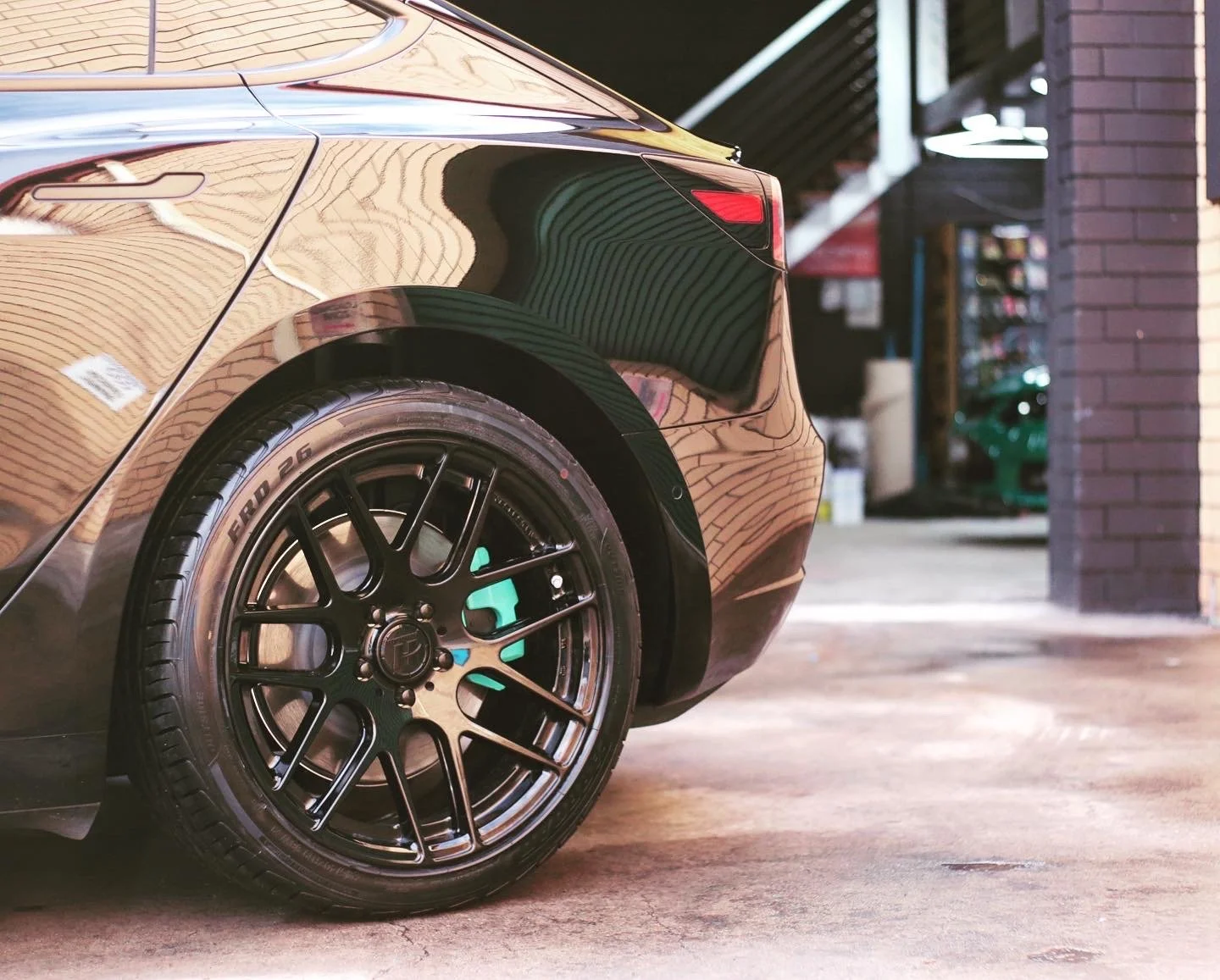 Close-up of the rear left side of a black sports car, showing the rear wheel with a black multi-spoke rim and low-profile tire, parked on a concrete surface in front of a building with brick walls and a staircase.