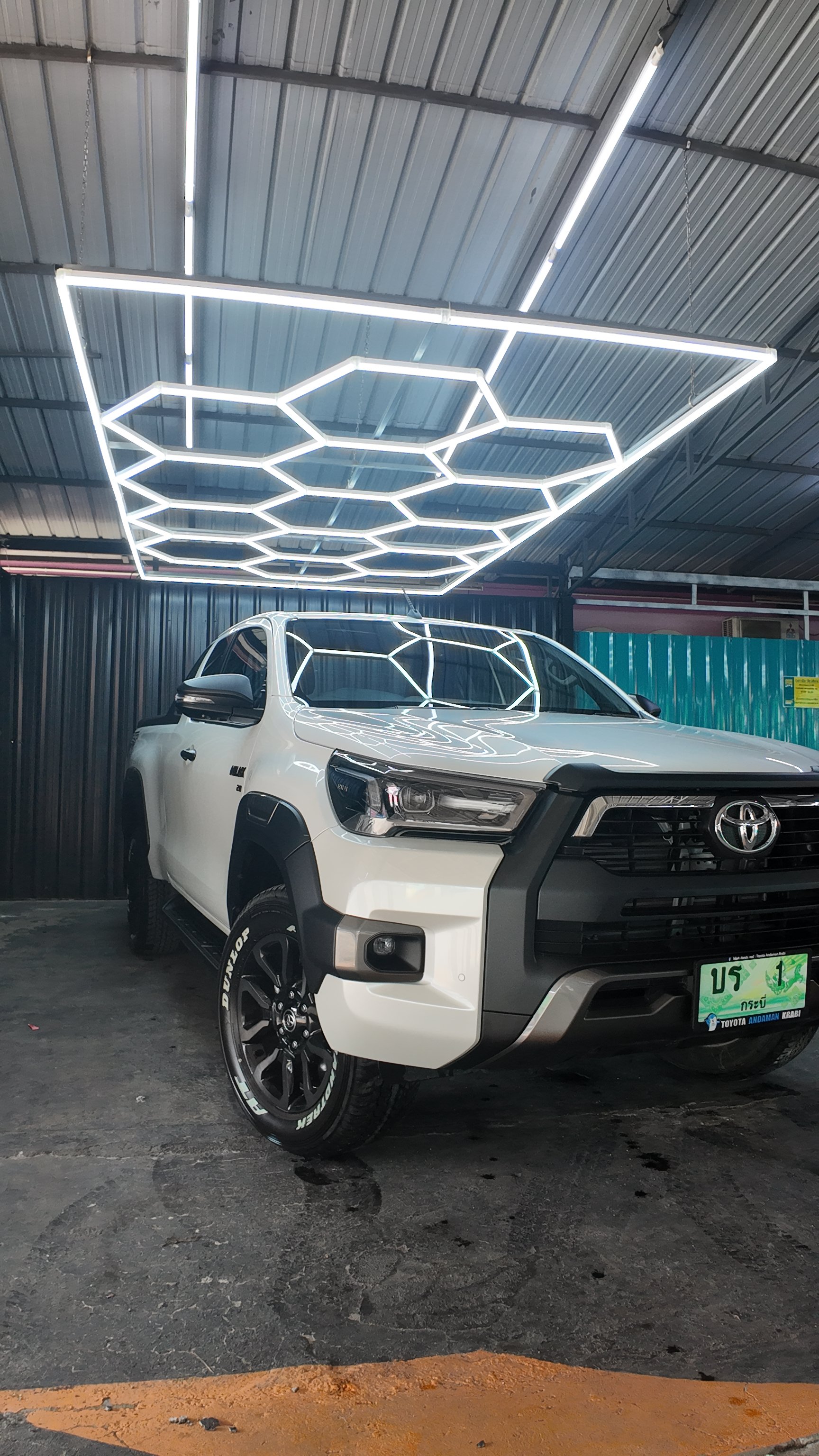 A white Toyota pickup truck parked under a metal roof with bright LED hexagon-shaped ceiling lights. The truck has black wheels, side mirrors, and a front grille. The ground is a dark asphalt surface with some visible dirt and wear.