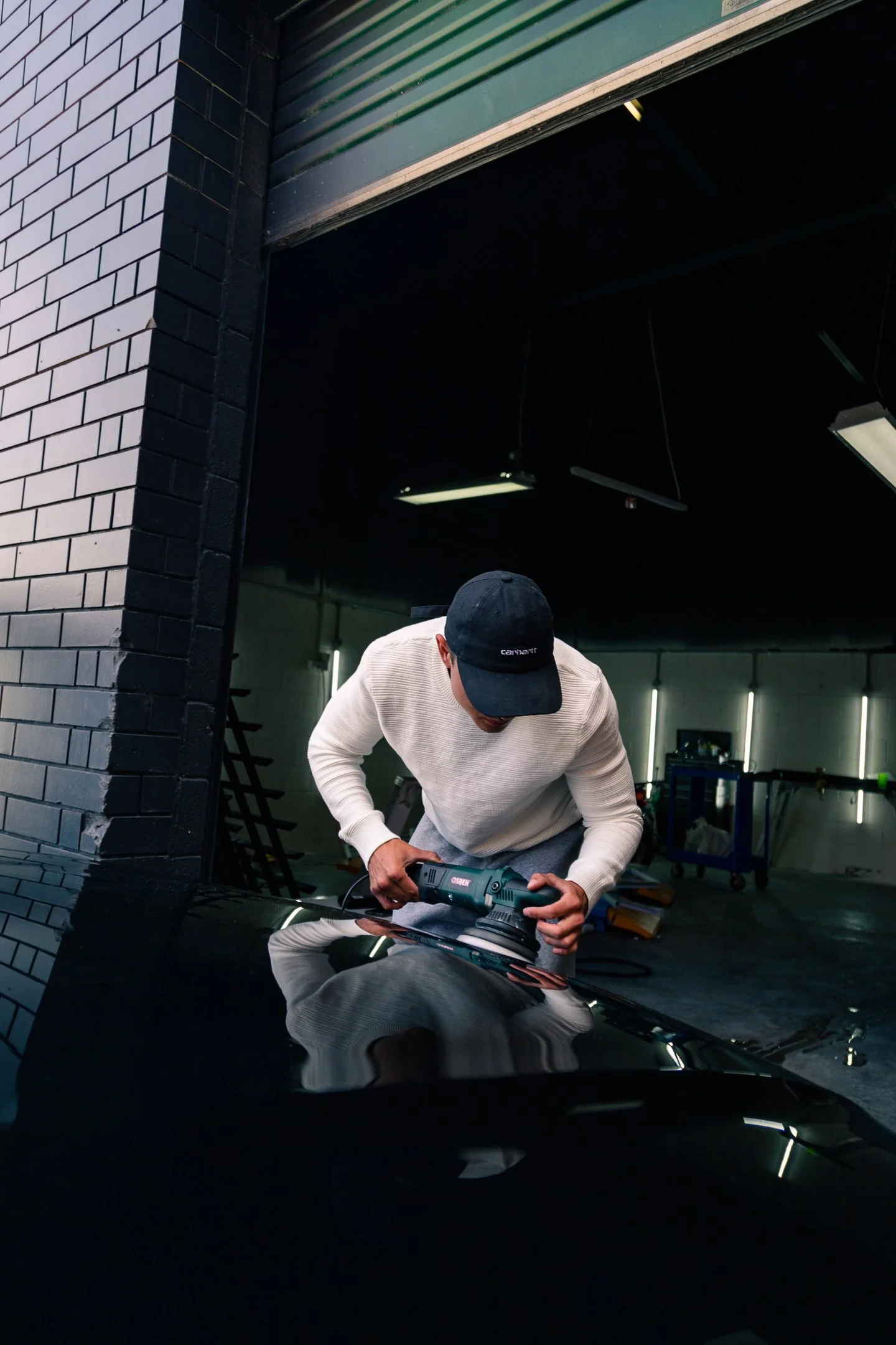 A person in a white sweater and black cap polishing a black car with a power buffer tool inside a garage, with the garage door open and fluorescent lights hanging from the ceiling.