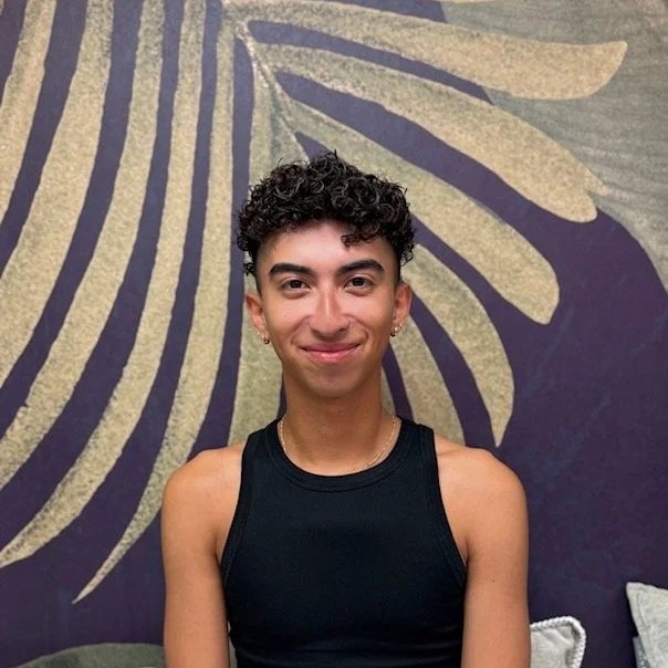 Young person with light skin wearing sunglasses, a gray New York Yankees cap, a light blue shirt, a white tank top, and jewelry, smiling with a beverage in hand against a white background.