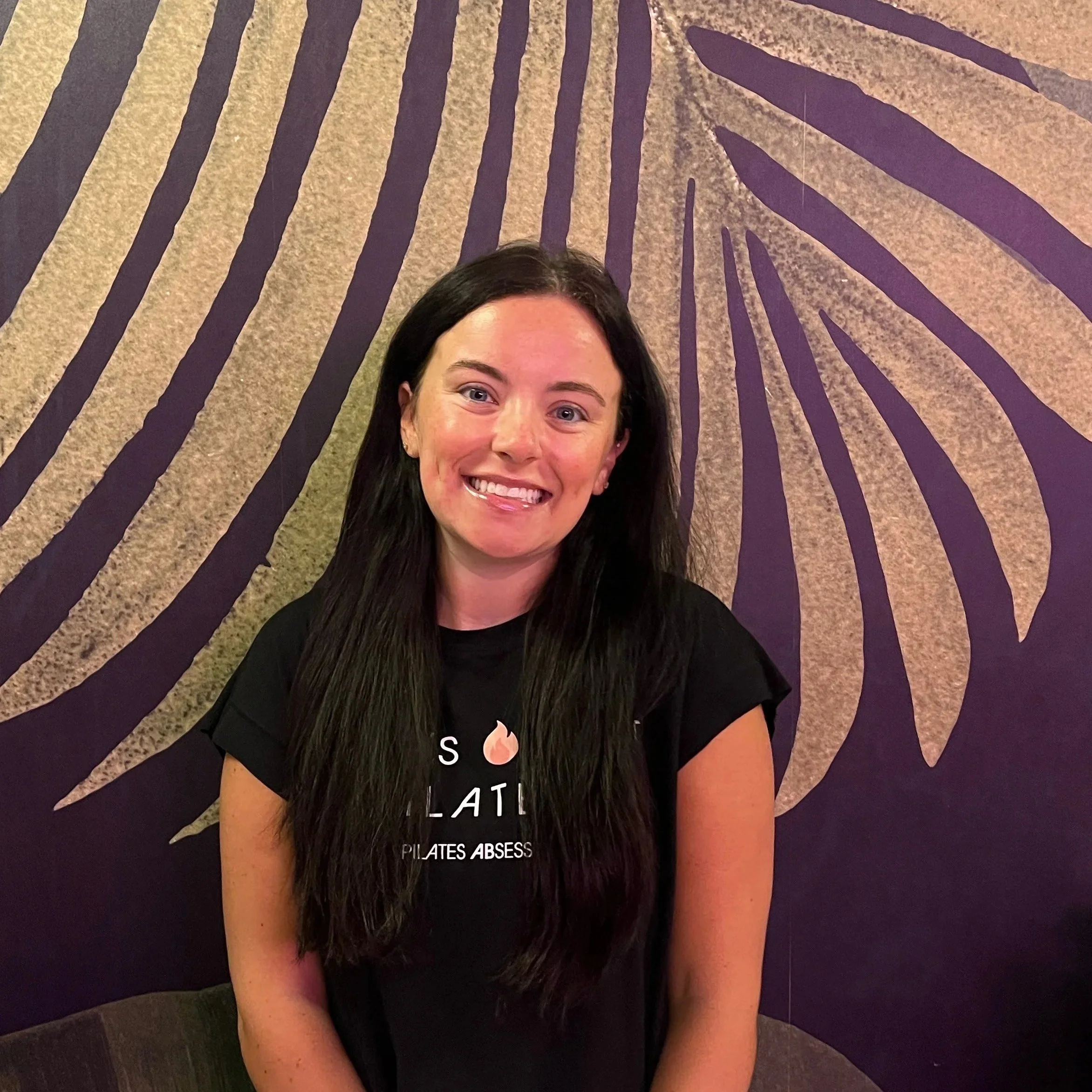 A woman wearing a wide-brimmed straw hat, a red swimsuit top, and a black hair tie, holding the brim of her hat with a slight smile against a bright white background.