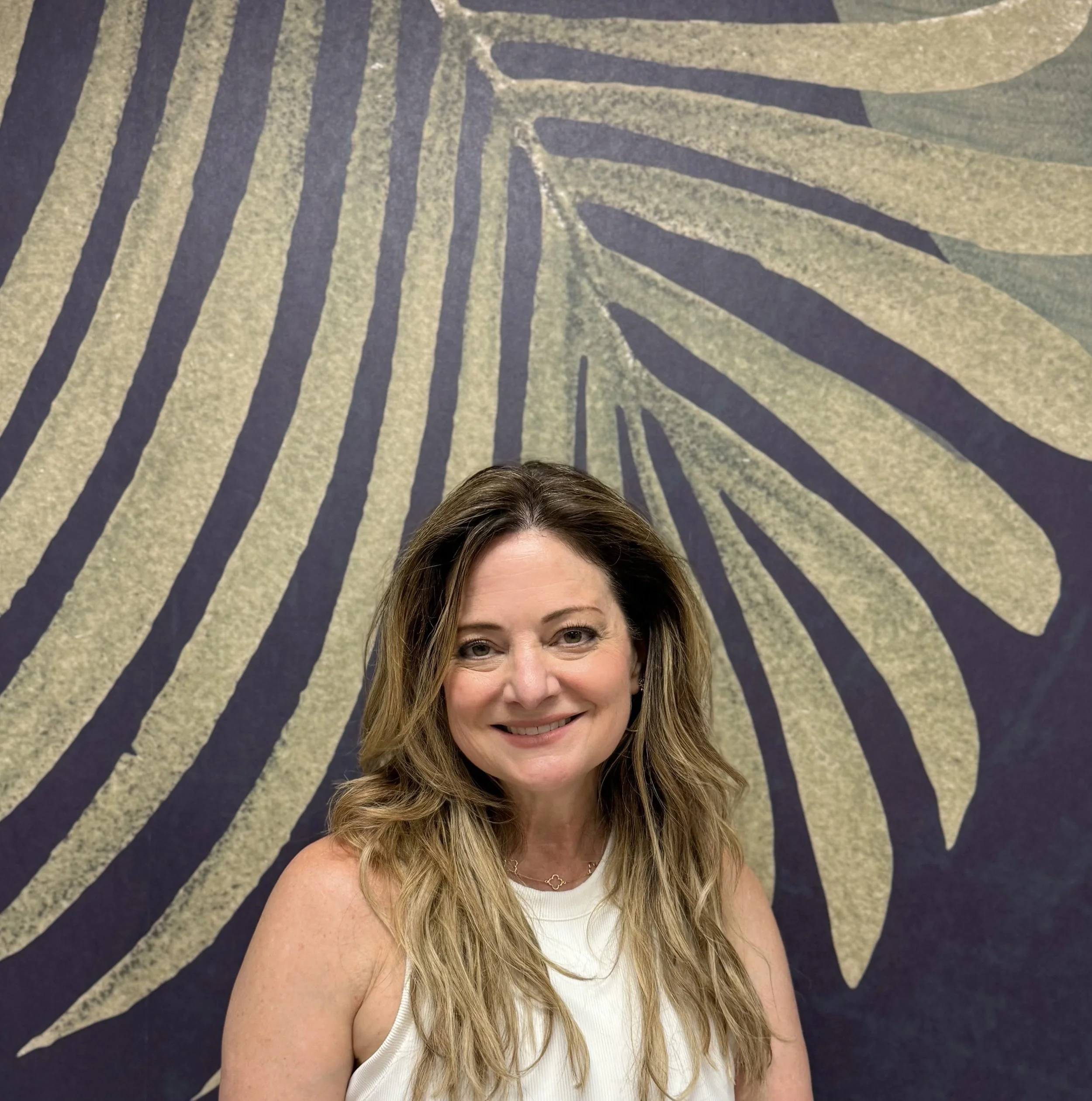 A woman with long wavy brown hair wearing a white sleeveless top, smiling at the camera, standing in front of a large abstract mural with black lines on a beige background.