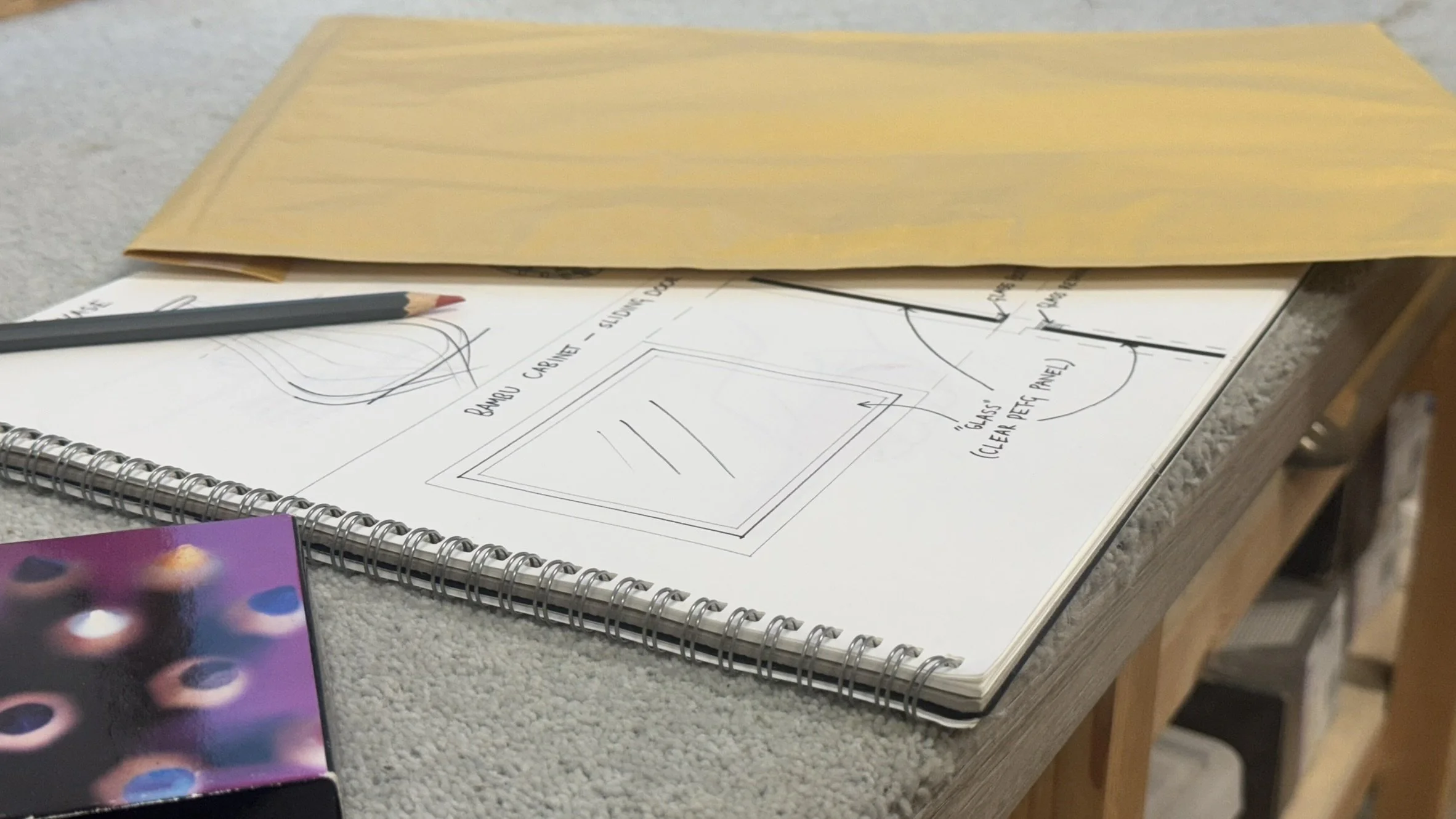Sketchbook with handwritten notes and sketches about cabinets and glass panel details, a black pencil, and a yellow envelope on a textured surface.