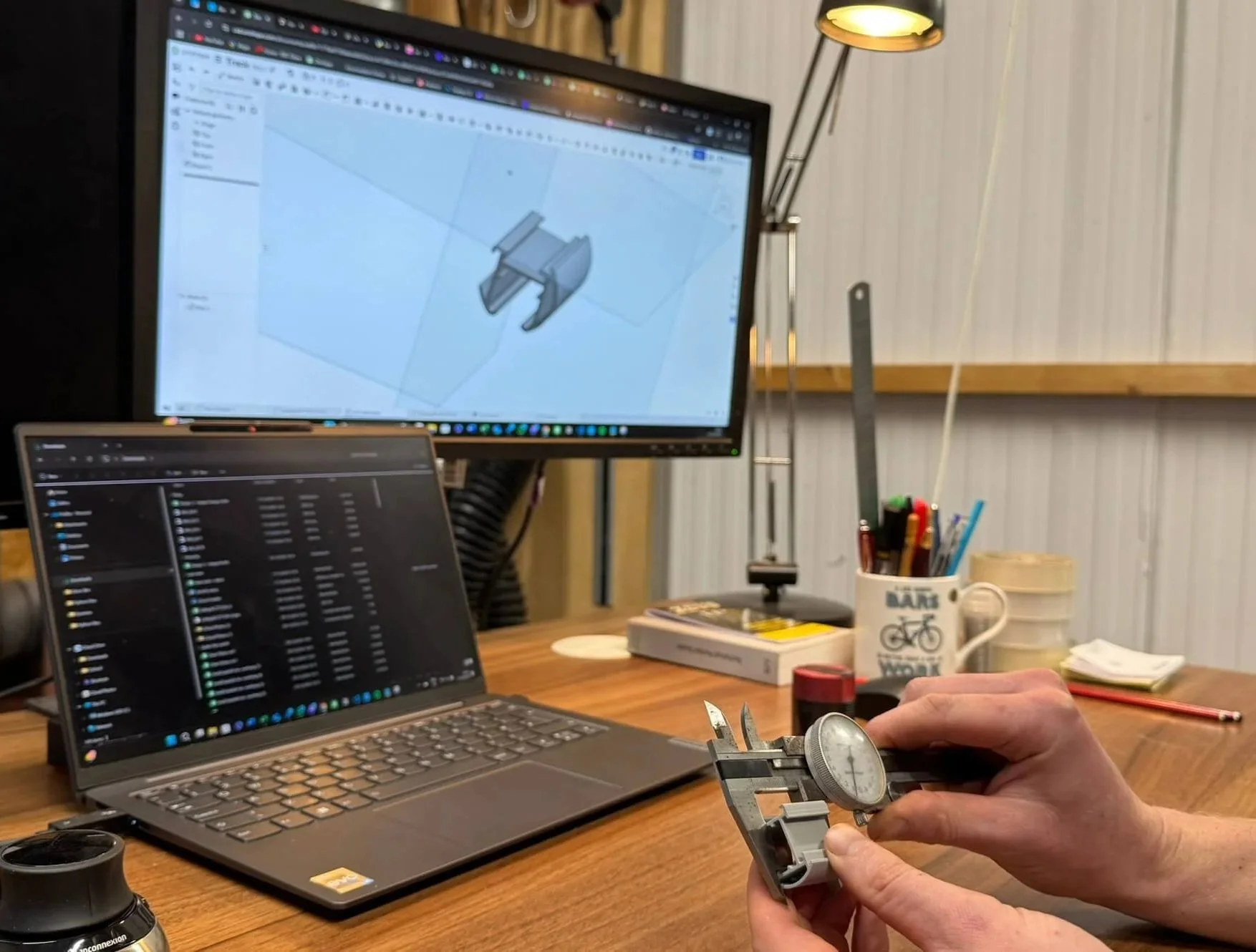 An engineer using vernier callipers to measure a 3D printed part infront of the design being shown on Fusion 360, on a pc screen
