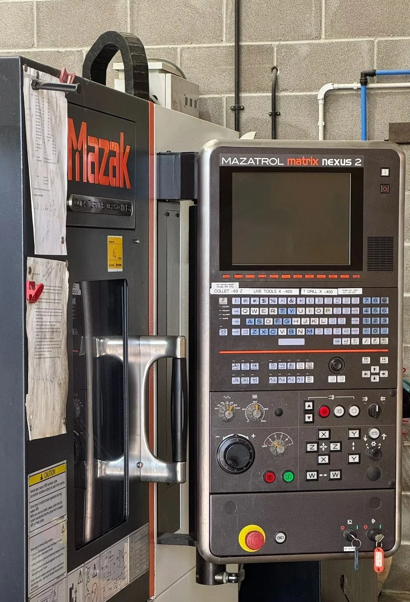 A Mazak CNC machine with a control panel, located in an industrial workshop with brick walls, pipes, and electrical wiring.