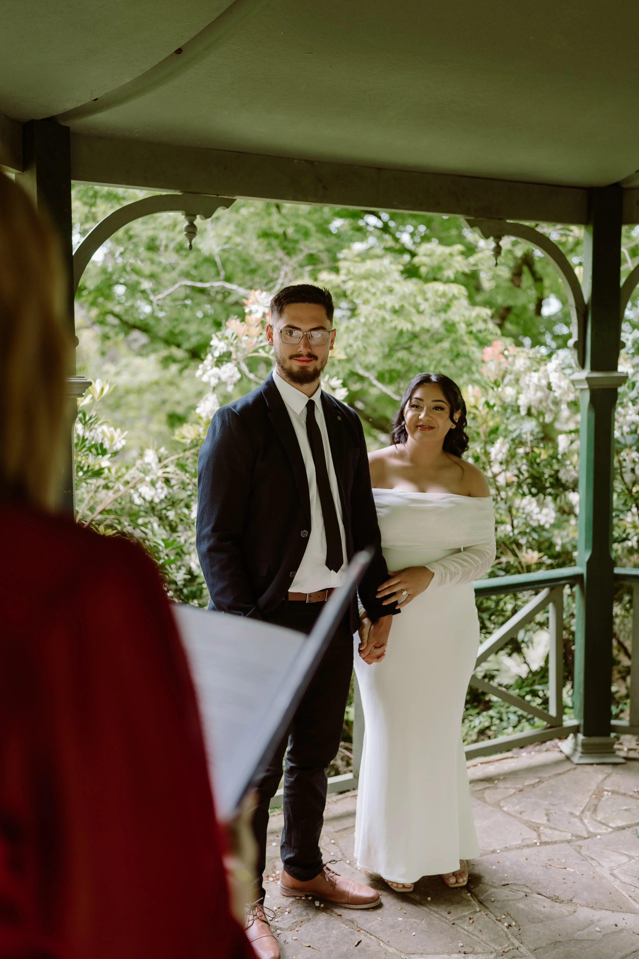 A wedding ceremony with a groom in a black suit and a bride in a white off-the-shoulder dress standing under a gazebo with lush greenery in the background.