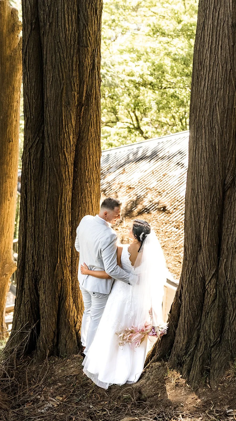 A bride and groom standing close together next to large trees, embracing each other outdoors during their wedding.