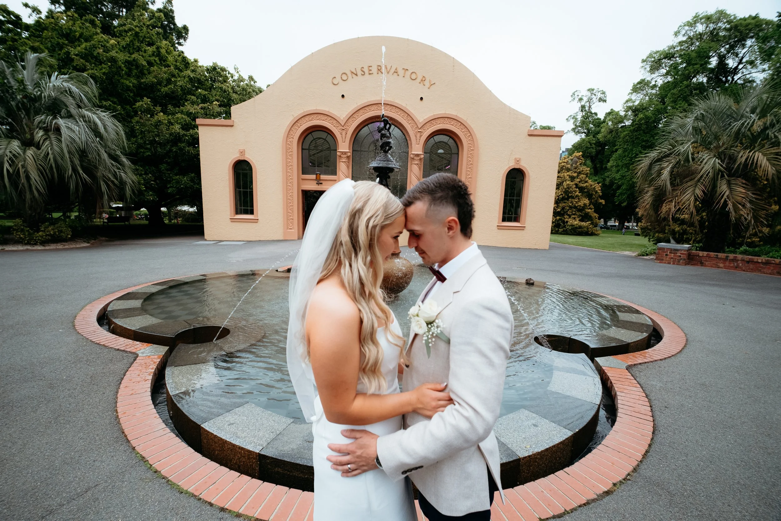 A bride and groom stand close together with foreheads touching in front of a small fountain outside a building labeled 'Conservatory'. The bride has long, wavy blonde hair and is wearing a white wedding dress and veil. The groom has short, dark hair and is dressed in a white suit with a dark bow tie and boutonniere.