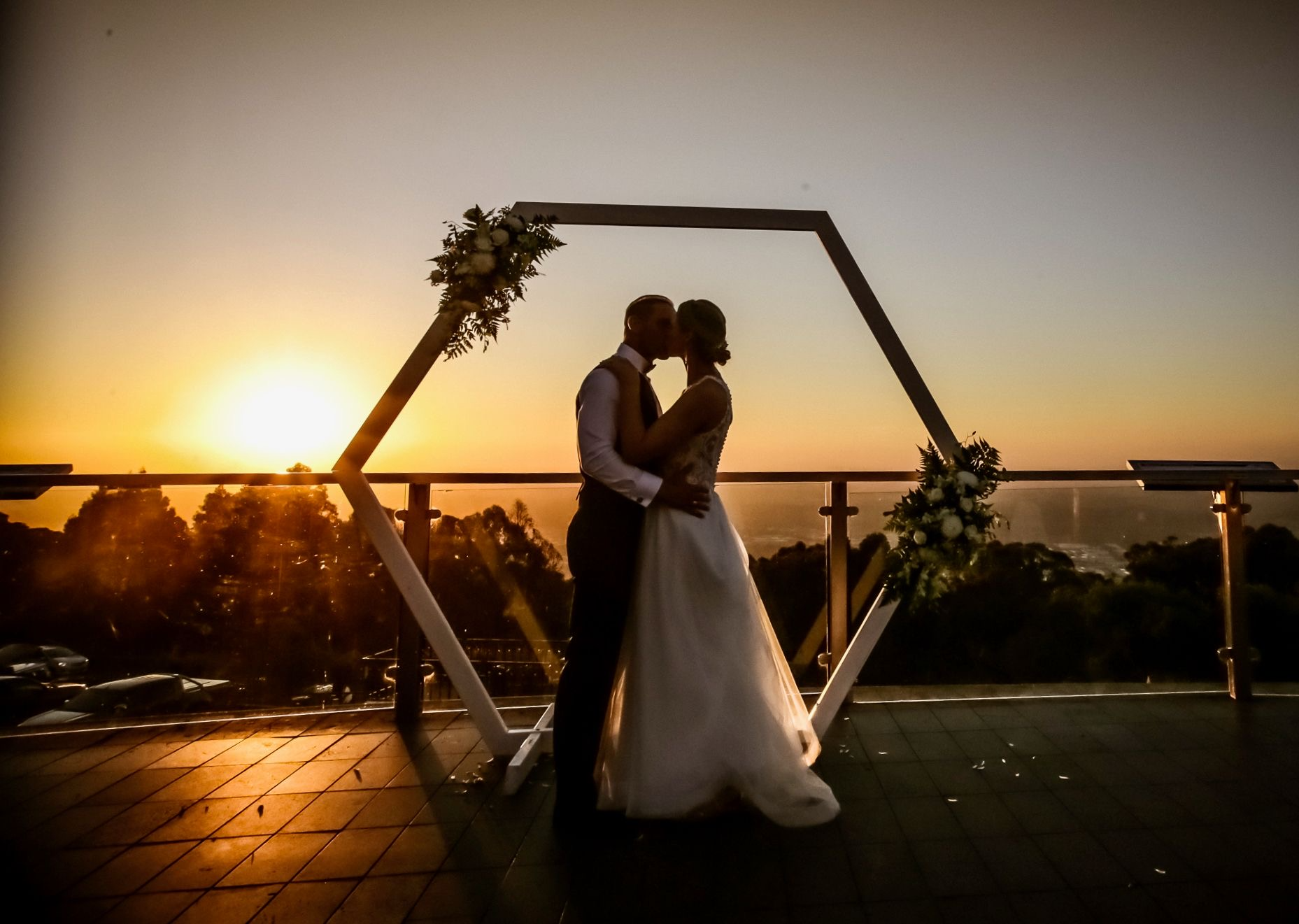Silhouette of a couple kissing during sunset at a wedding ceremony, standing underneath a geometric arch decorated with flowers, on a terrace with a railing and trees in the background.