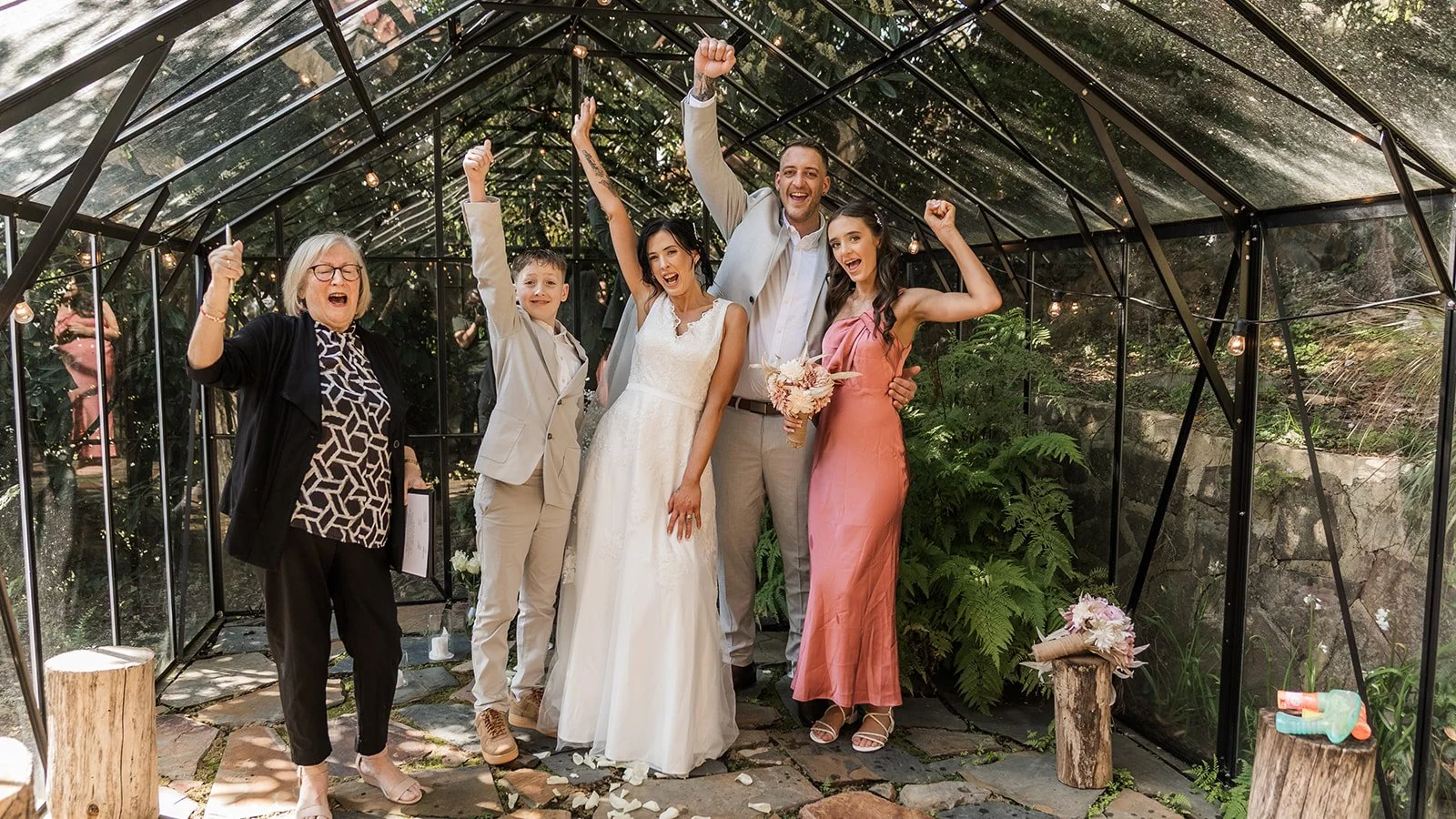 A group of people celebrating a wedding inside a glass greenhouse, with two women, a man, a boy, and a girl, cheering and smiling, some with raised fists, and flower arrangements on logs.