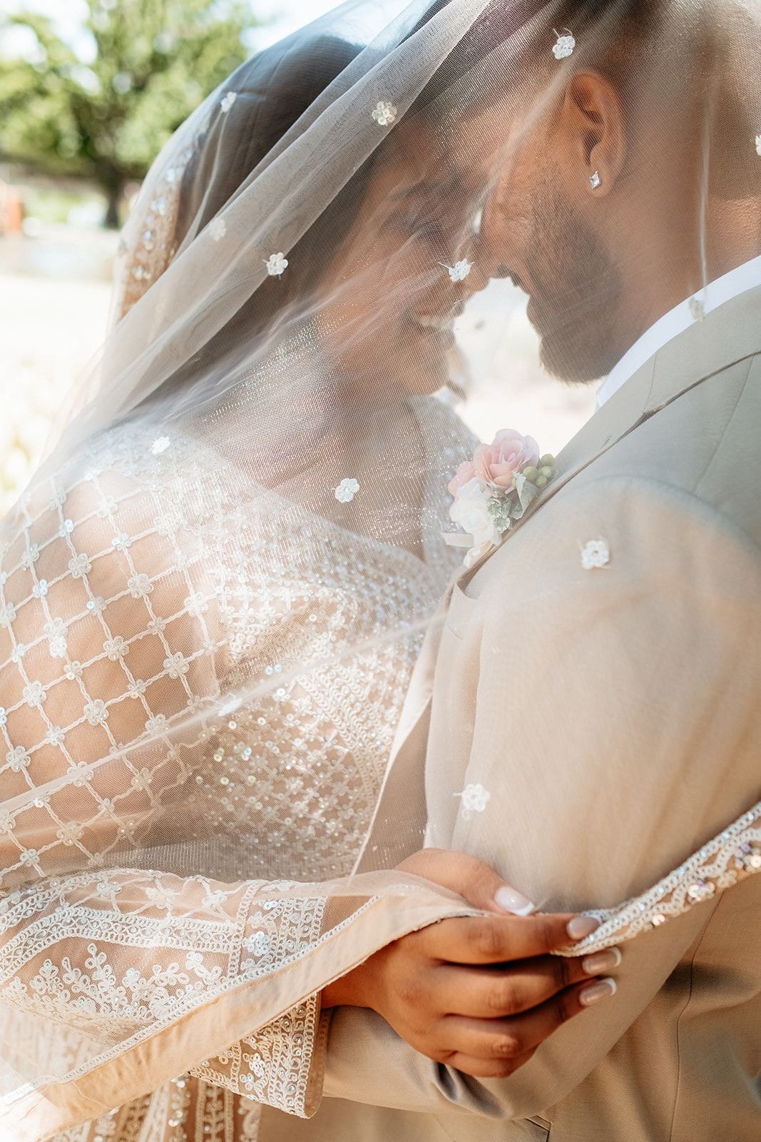 A bride and groom are close together, touching foreheads and smiling beneath a sheer veil with floral embellishments. The bride wears a lace wedding dress with detailed embroidery and the groom is in a beige suit.