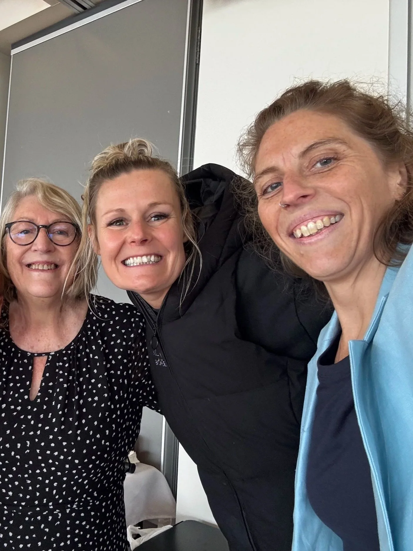 Three women smiling and taking a selfie together in an indoor setting, with a gray door or partition in the background.