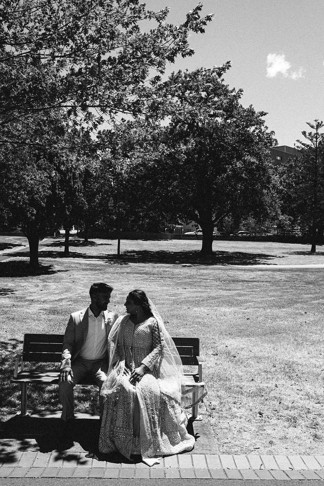 A black-and-white photo of a couple sitting on a park bench, with trees and a grassy area in the background. The woman is wearing a wedding dress with a veil, and the man is dressed in a suit.