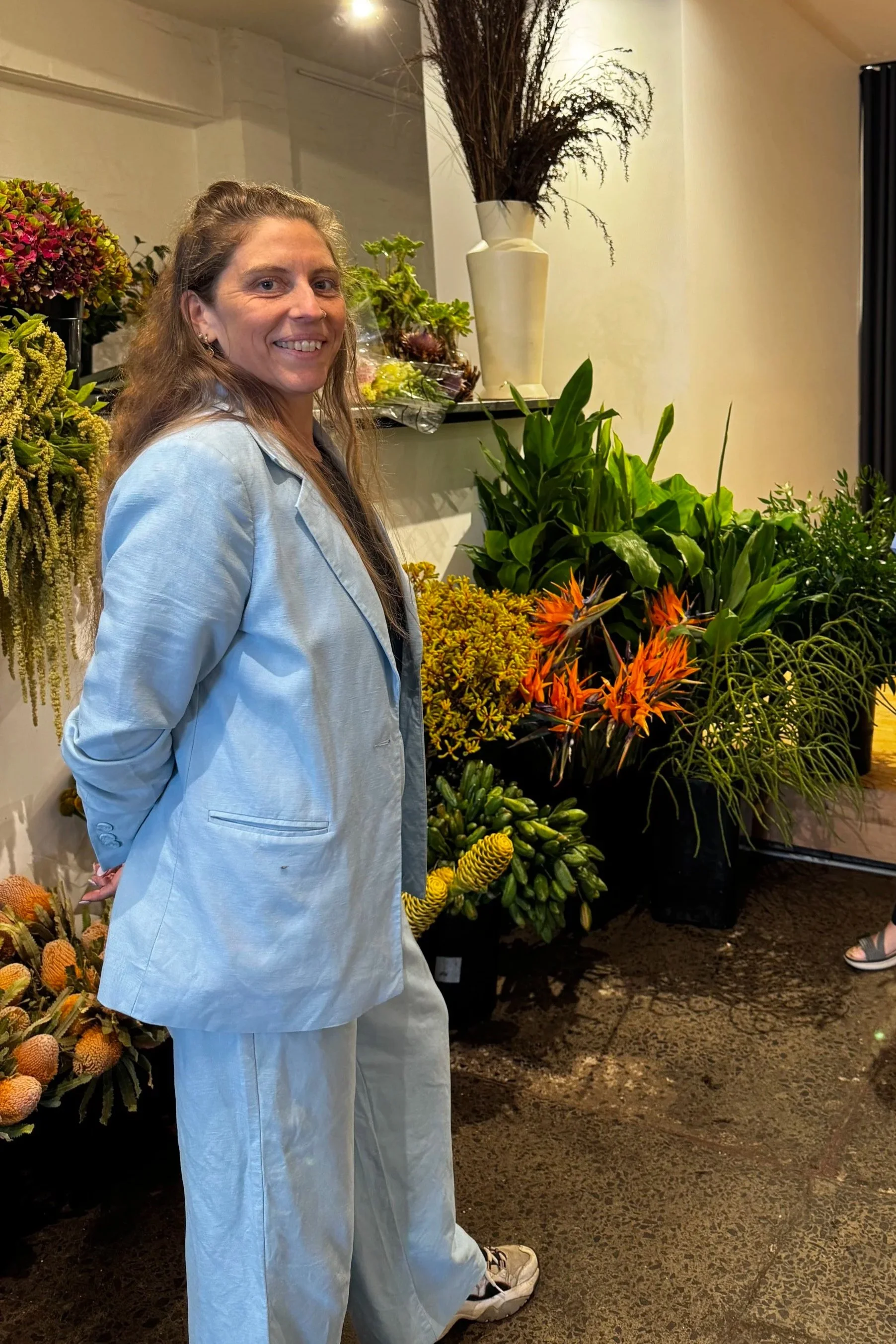A woman in a light blue suit standing in front of a display of various colorful flowers and plants indoors.