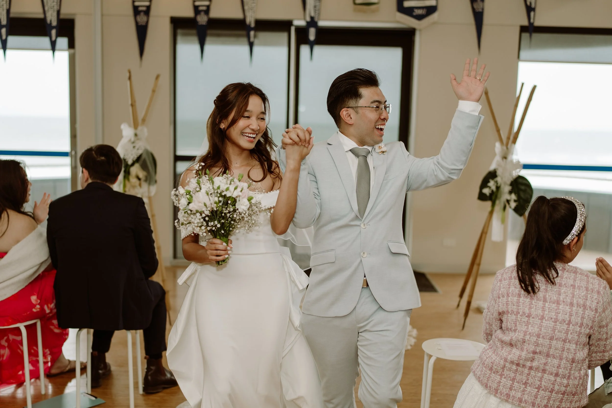 A wedding reception with a smiling bride and groom walking hand-in-hand, celebrating indoors with friends and family, near windows overlooking the ocean.